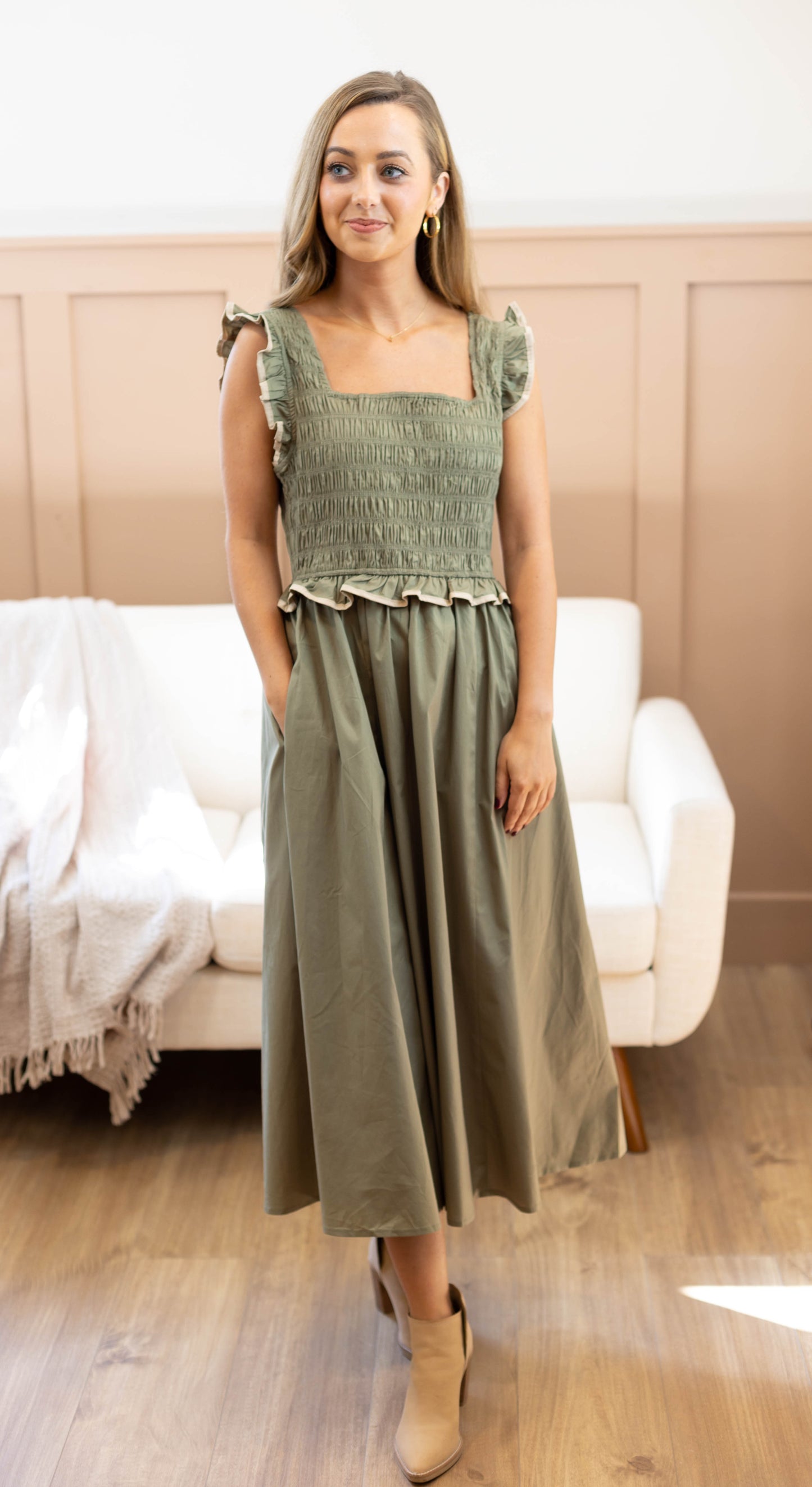Someone wearing the Clara Smocked Midi by Dogwood Cloth in fir-tree green, featuring ruffle sleeves, stands in a room with a white couch and wooden floor.