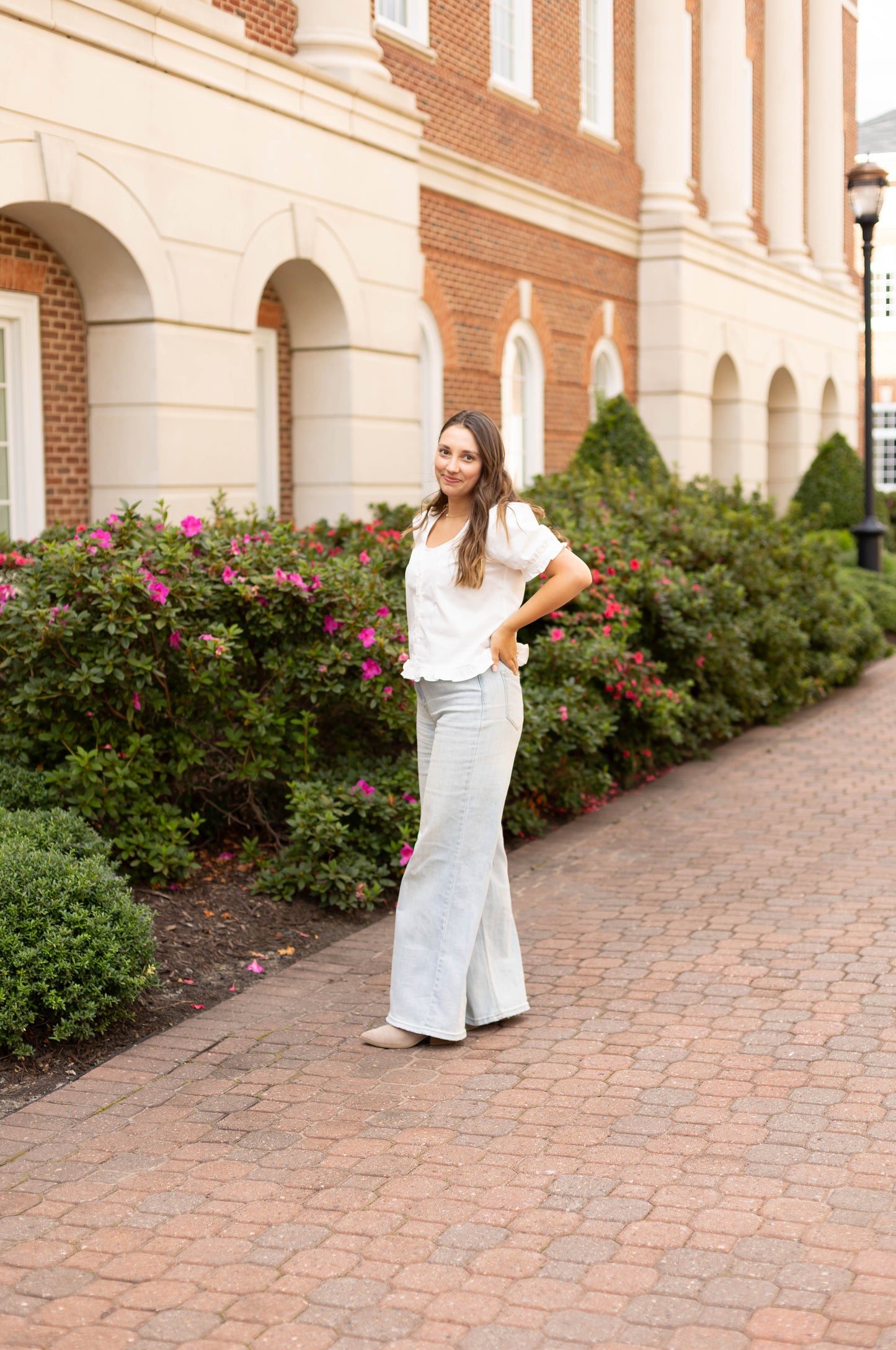 A woman stands on a brick pathway in front of bushes and archways, wearing the Dogwood Cloth Cory Top, a white cotton puff sleeve blouse, with light gray pants.