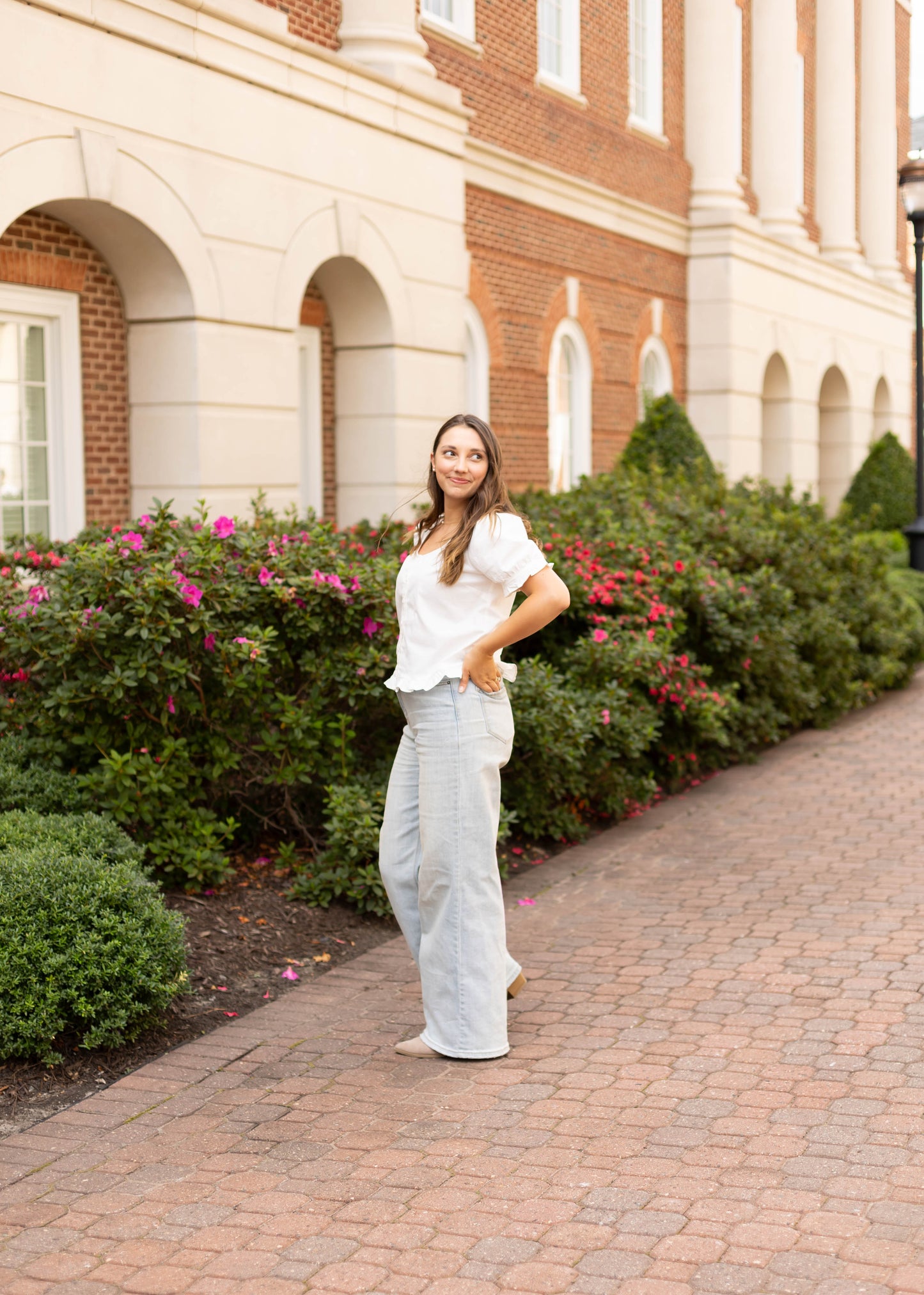 A woman wearing the Dogwood Cloth Cory Top and light gray pants stands on a brick walkway beside green bushes and pink flowers in front of a brick building.