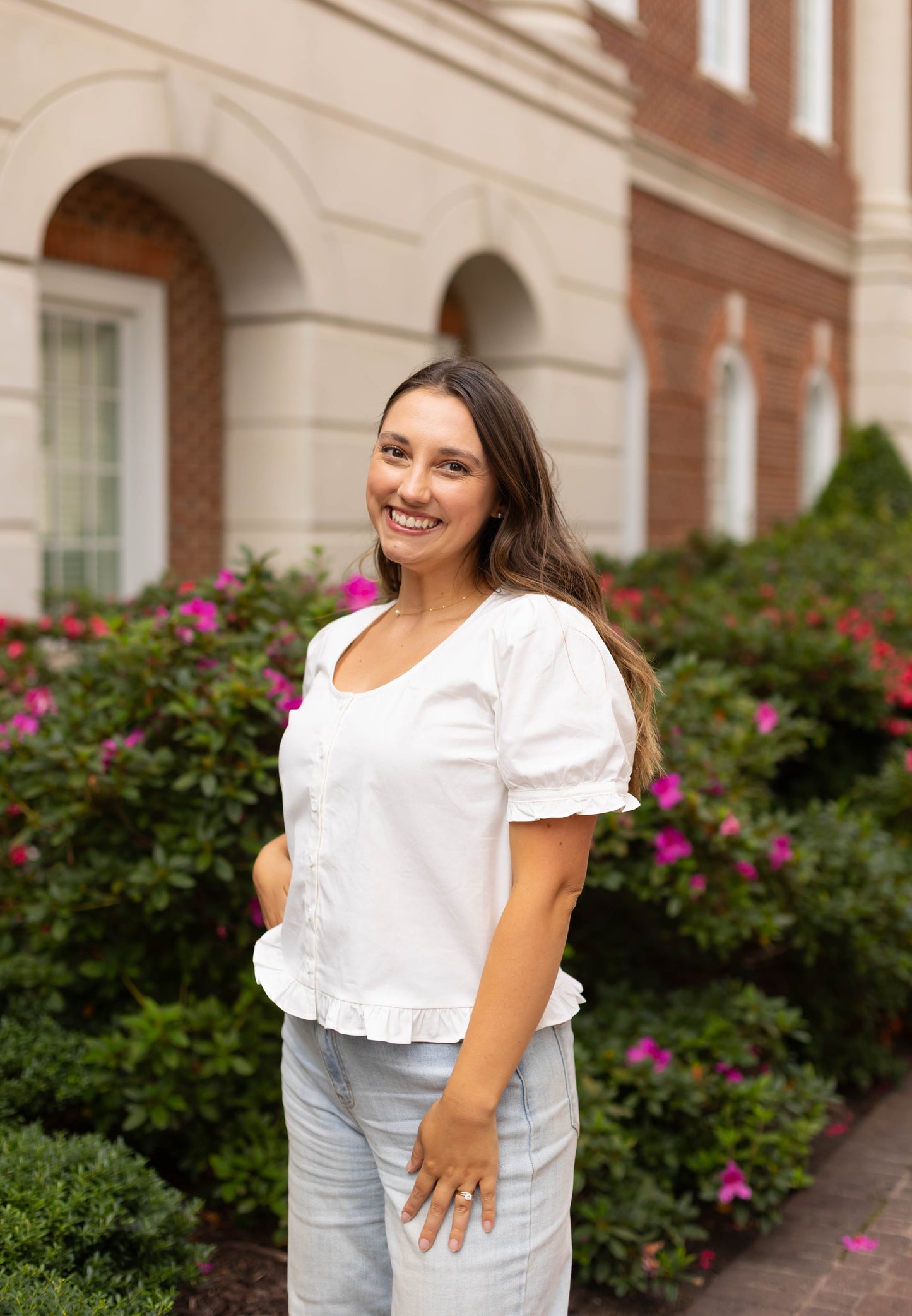 A woman smiles outdoors in front of a brick building and blooming shrubs, wearing the Dogwood Cloth Cory Top, a white cotton blouse, paired with light blue pants.