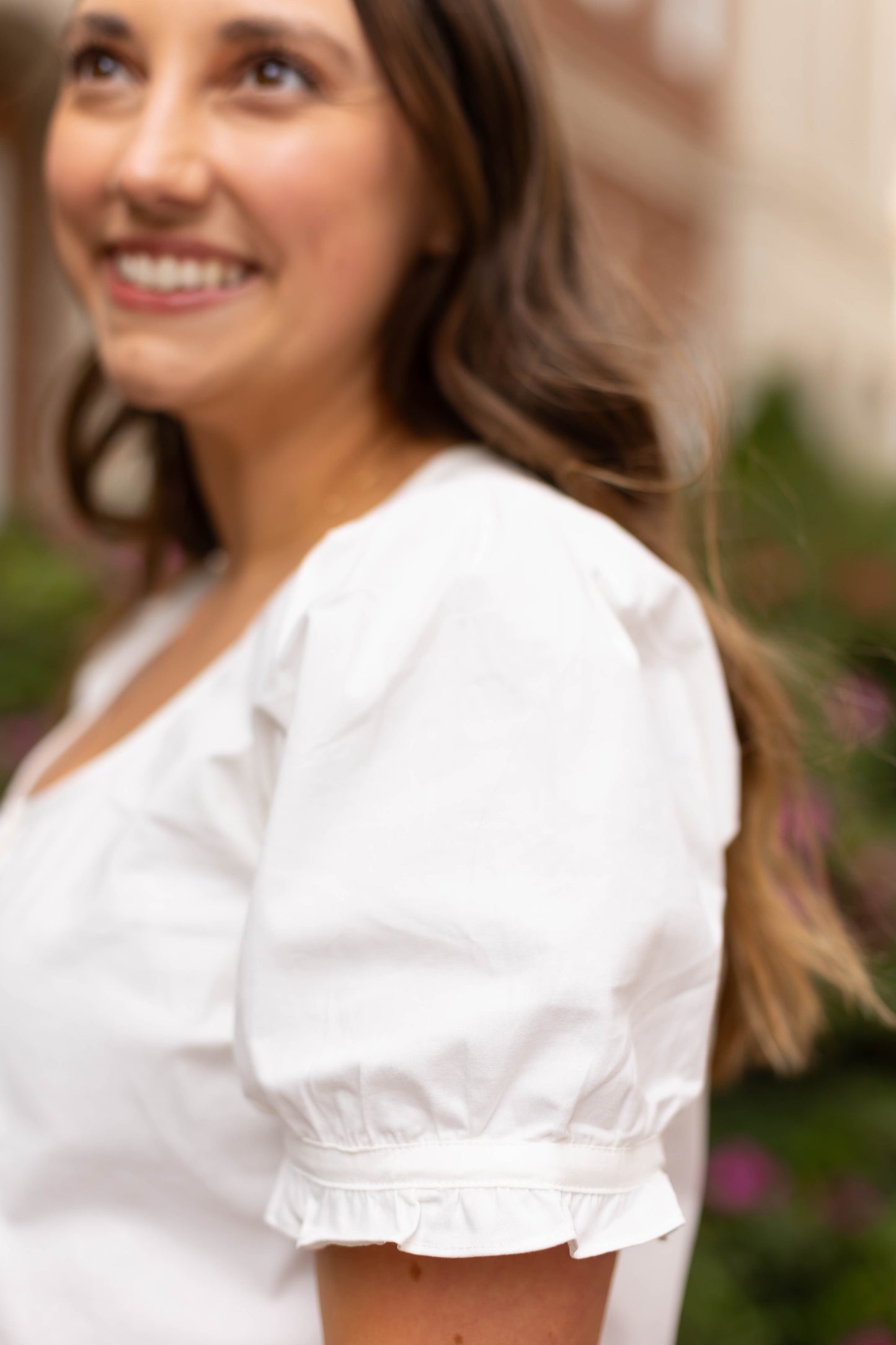 A woman with long brown hair is smiling outdoors, wearing the white Cory Top by Dogwood Cloth; greenery and buildings are softly blurred in the background.