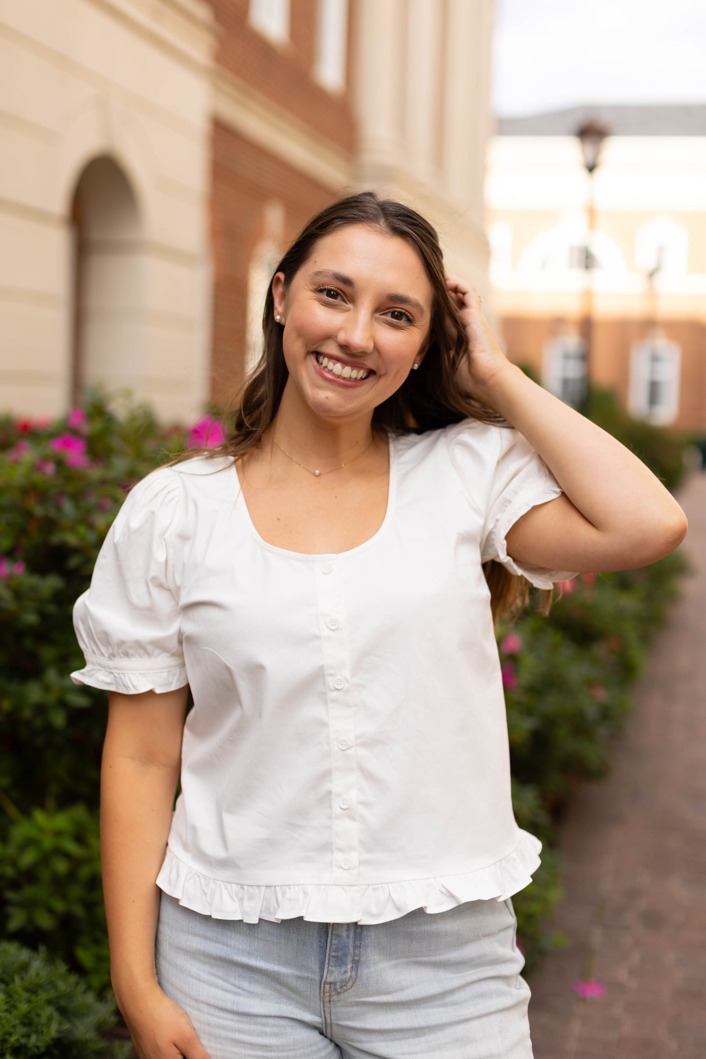 A young woman with long brown hair stands outdoors on a brick path, smiling at the camera with one hand in her hair, wearing the Cory Top by Dogwood Cloth and light jeans.
