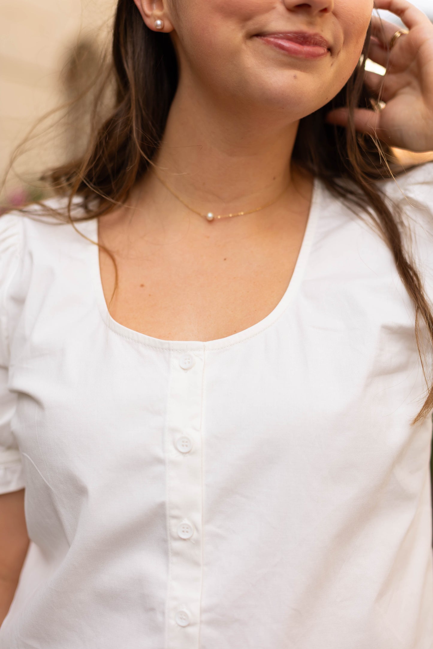 Close-up of a woman in the Cory Top by Dogwood Cloth—a white cotton blouse with puff sleeves—paired with a delicate gold necklace featuring a small pearl pendant, as she touches her hair.