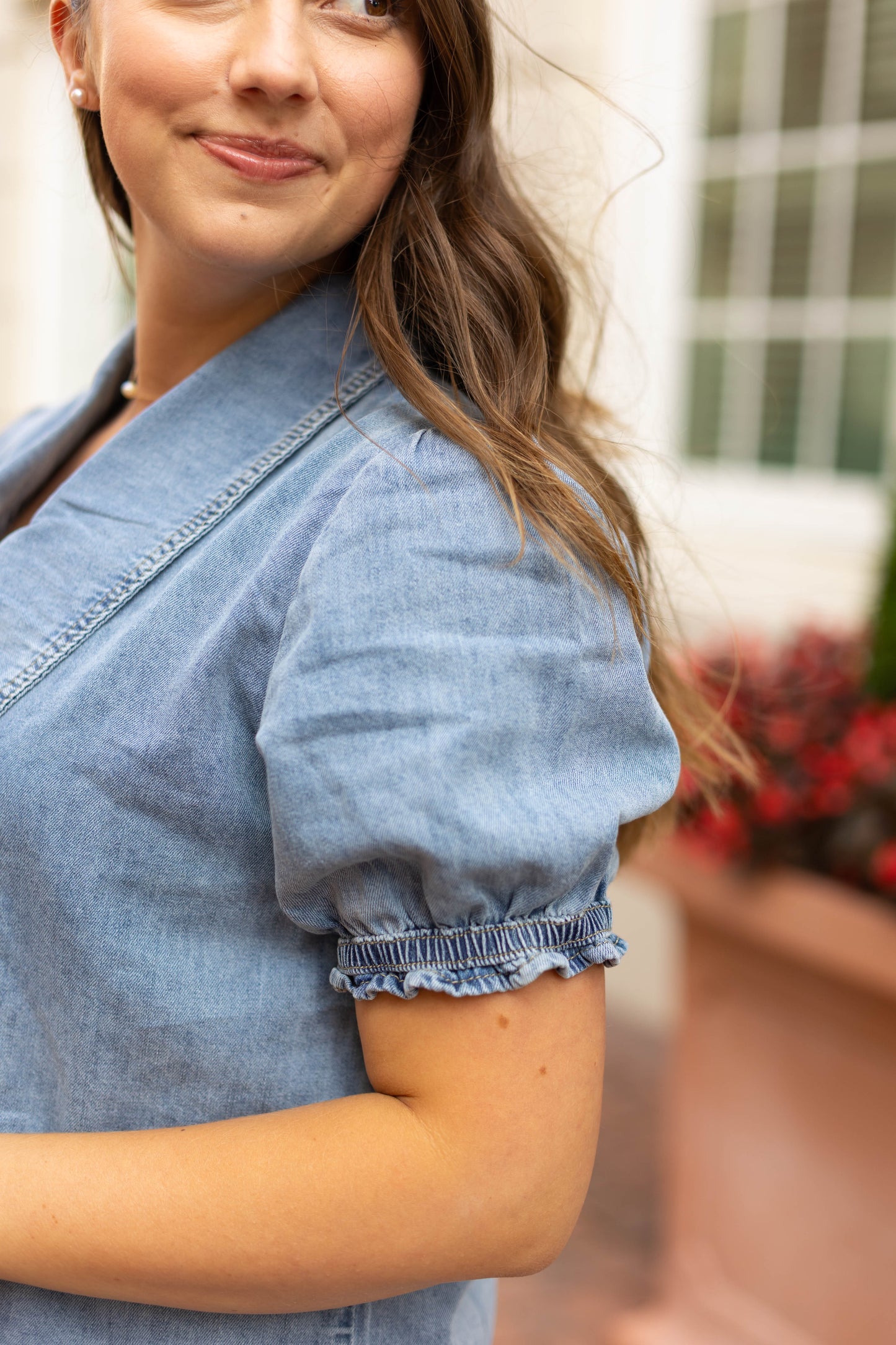 A woman with long brown hair wears the Dogwood Cloth Jessa Jean Top, a light blue button-up blouse with puff sleeves, while standing outdoors near a building and a planter filled with red flowers.