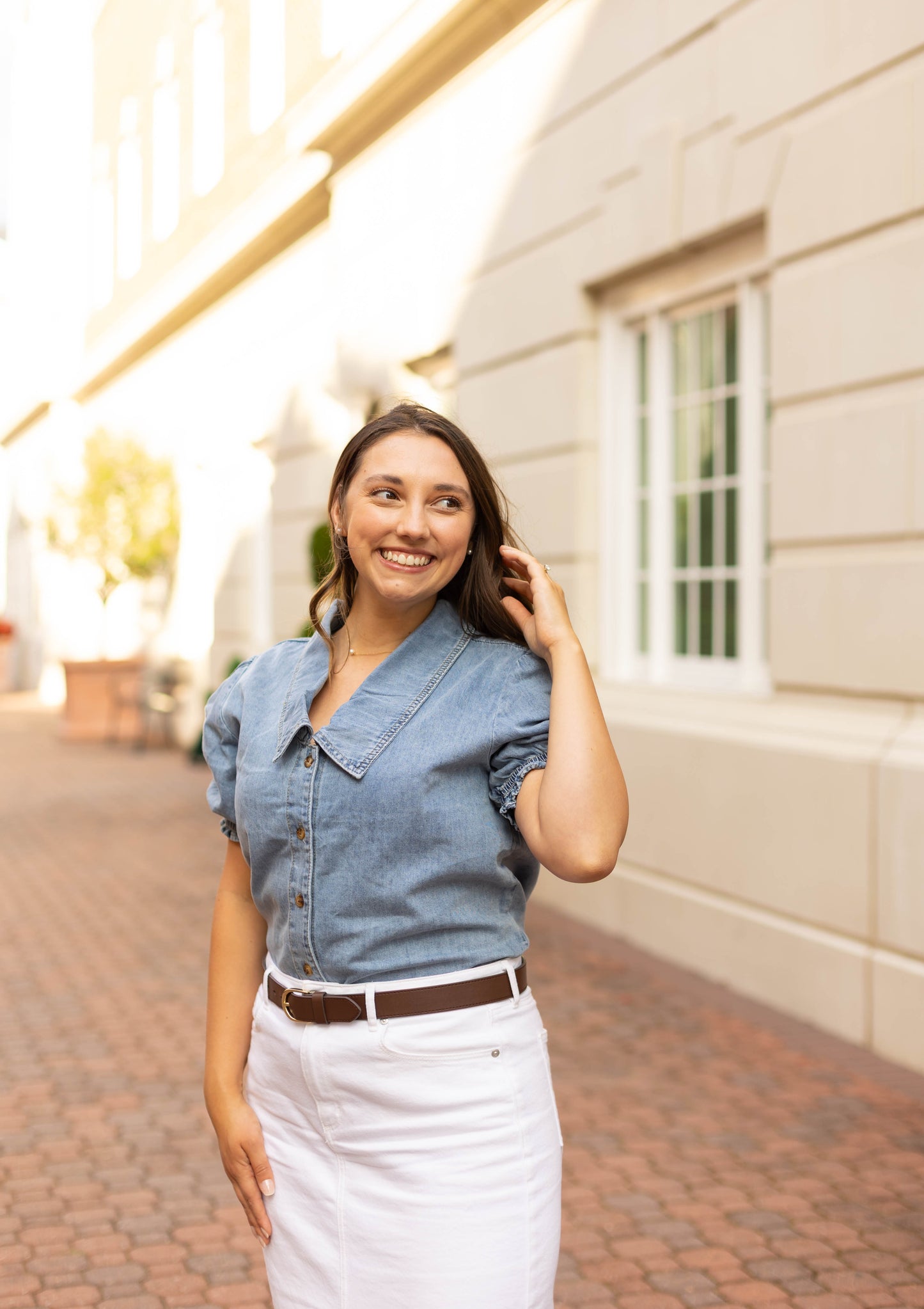 A woman stands outdoors on a brick walkway, smiling and touching her hair, wearing the Dogwood Cloth Jessa Jean Top with puff sleeves and a white skirt, with a building and windows in the background.