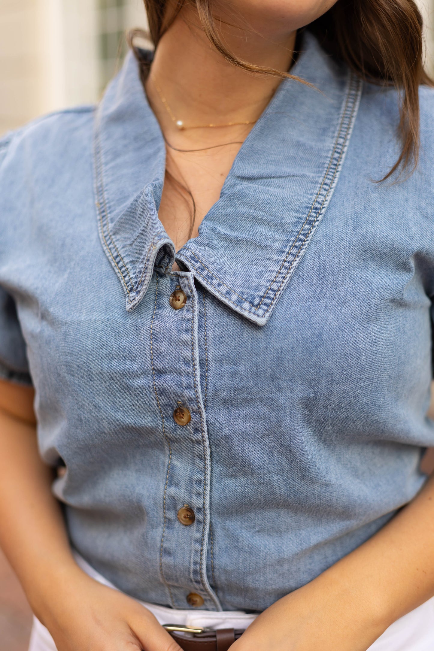 A woman wears the Dogwood Cloth Jessa Jean Top, a blue denim button-up blouse with a wide collar and puff sleeves, paired with a delicate gold necklace.