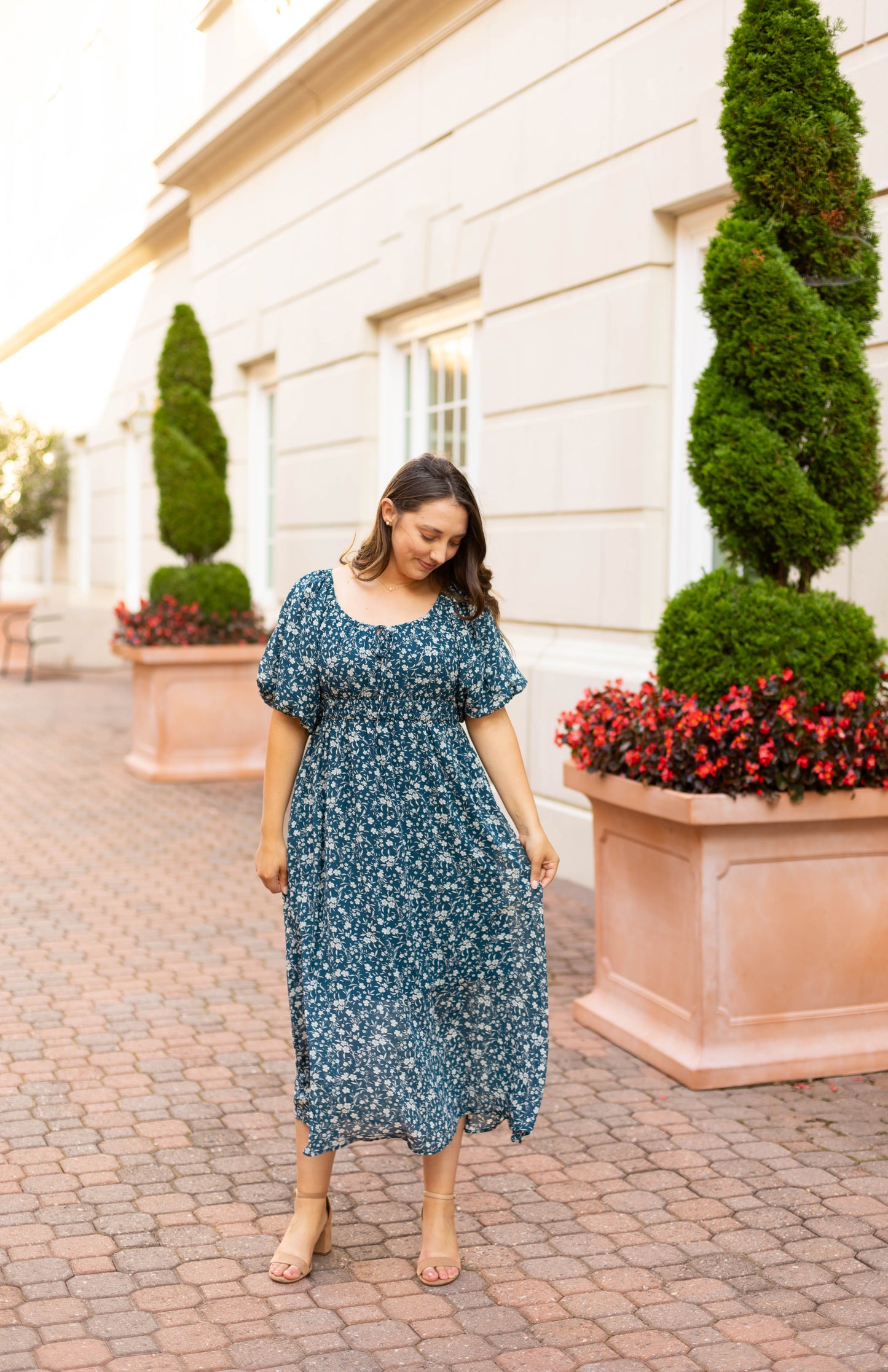 A woman wearing the Dogwood Cloth Aubrey Midi Dress stands on a paved walkway between large planters with spiral-trimmed bushes and red flowers, near a light-colored building.