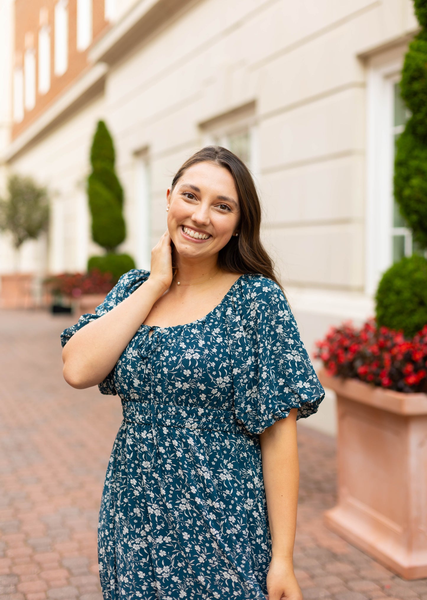 A woman wearing the Dogwood Cloth Aubrey Midi Dress smiles at the camera while standing on a brick pathway beside a building with potted plants.