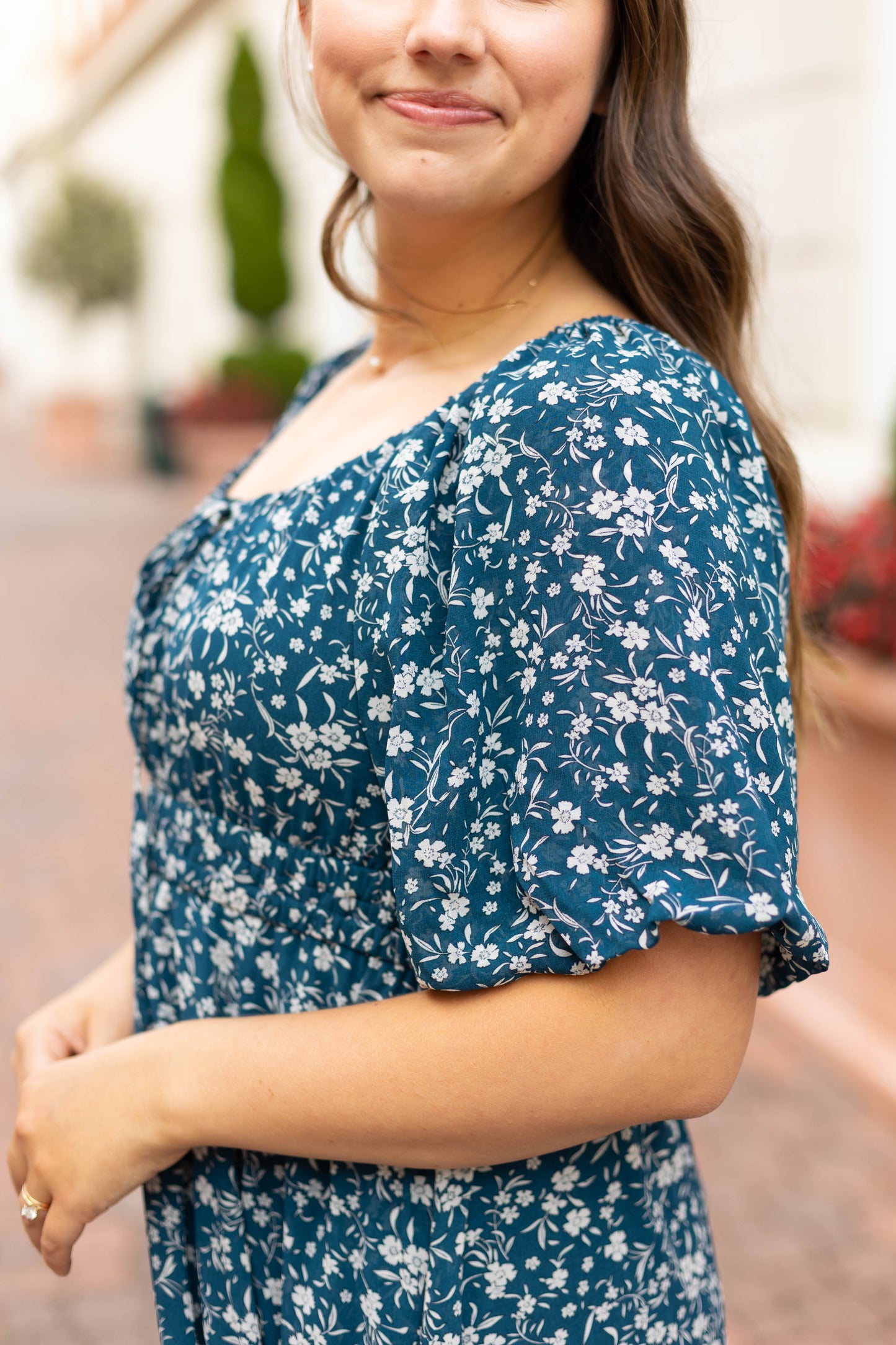 A woman in the Dogwood Cloth Aubrey Midi Dress with an empire waist stands outdoors on a brick pathway, her hands lightly clasped, with greenery and buildings softly blurred in the background.
