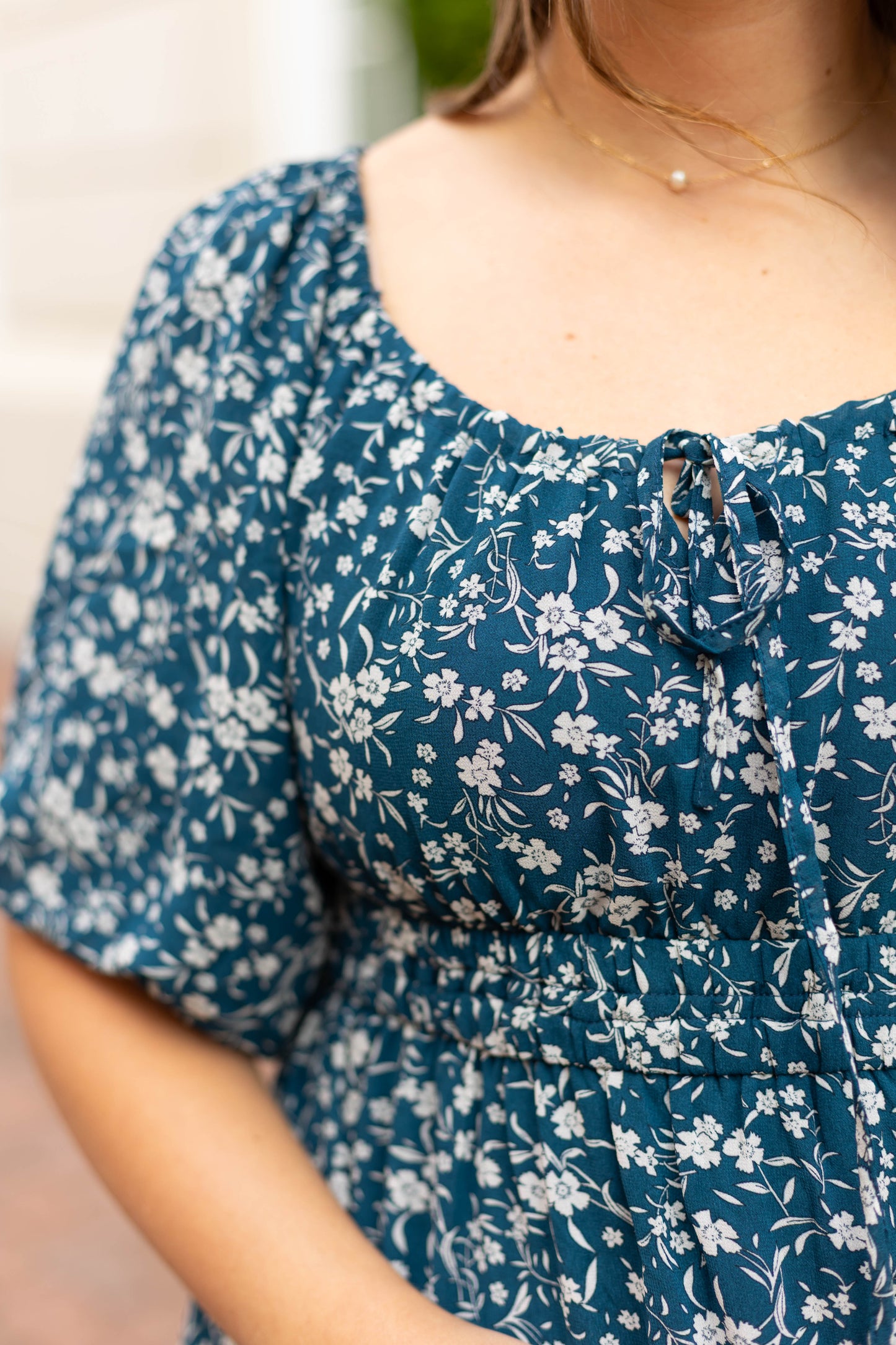 A woman wearing the Dogwood Cloth Aubrey Midi Dress, a blue midi dress with white floral pattern, puffed sleeves, and an empire waist, shown from shoulders to waist.