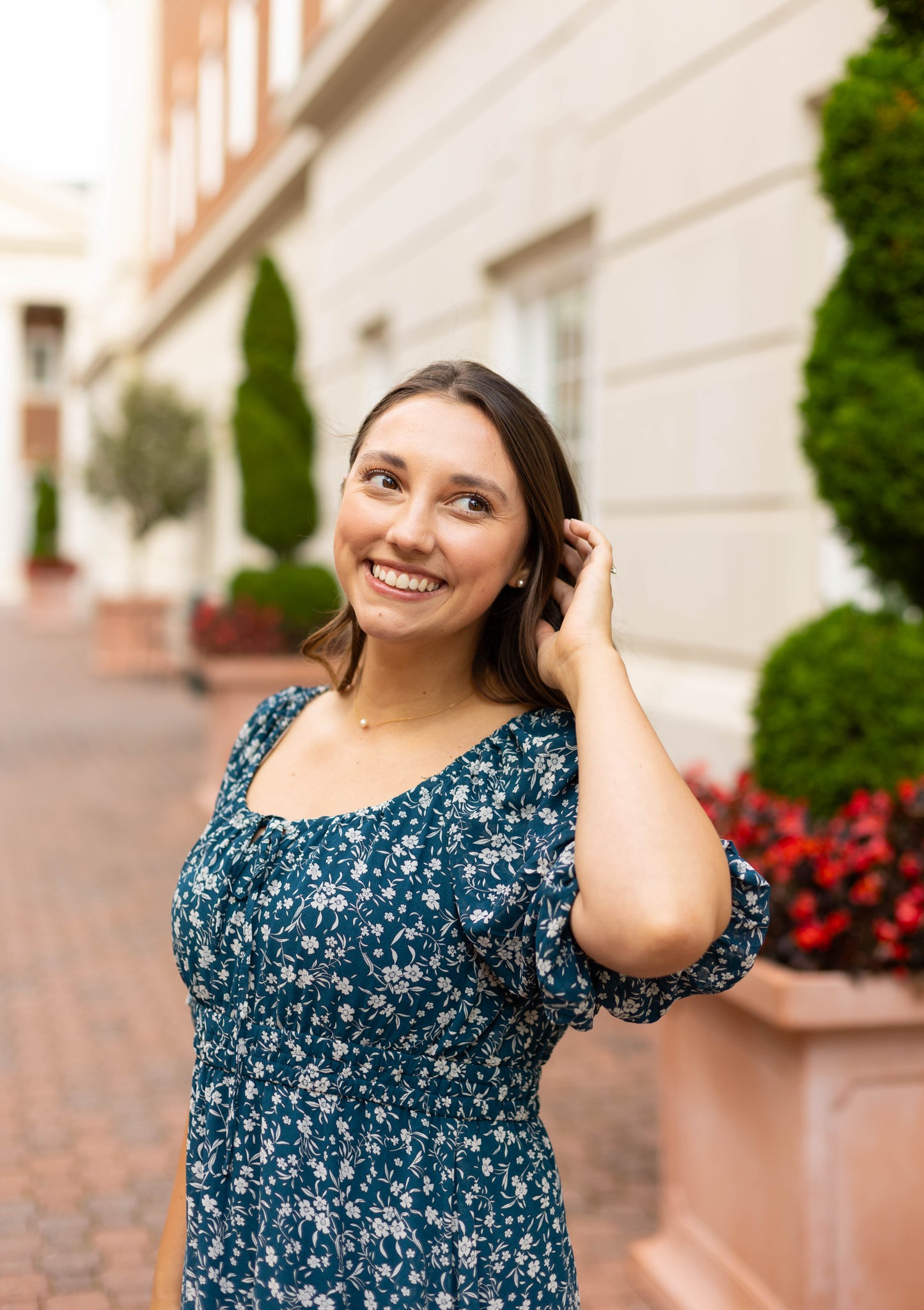 Wearing the Dogwood Cloth Aubrey Midi Dress with an empire waist, a woman stands on a brick walkway, smiling and touching her hair amid green foliage and red flowers.