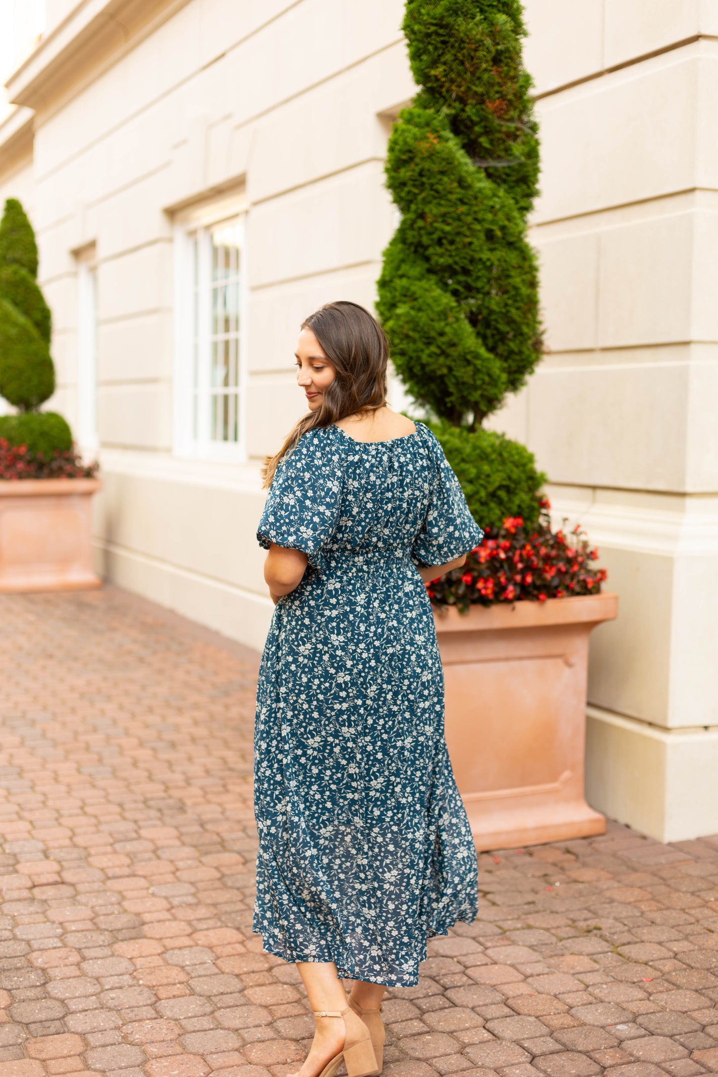 A woman in the elegant blue floral Aubrey Midi Dress by Dogwood Cloth, featuring an empire waist, walks along a brick path beside a light building with large planters and manicured shrubs.