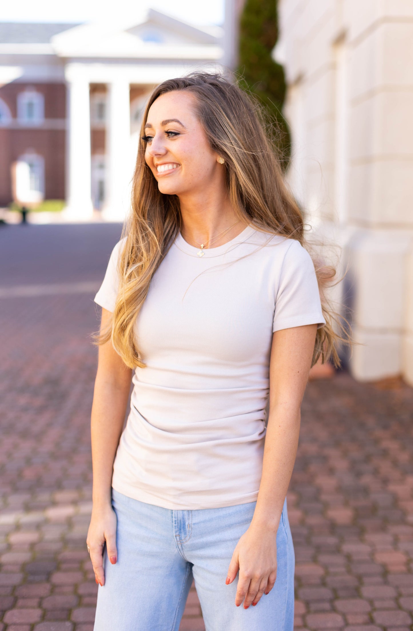 A woman with long hair, wearing a Dogwood Cloth's Marie Everyday Top and jeans, stands outdoors on a brick walkway, smiling and looking to the side.