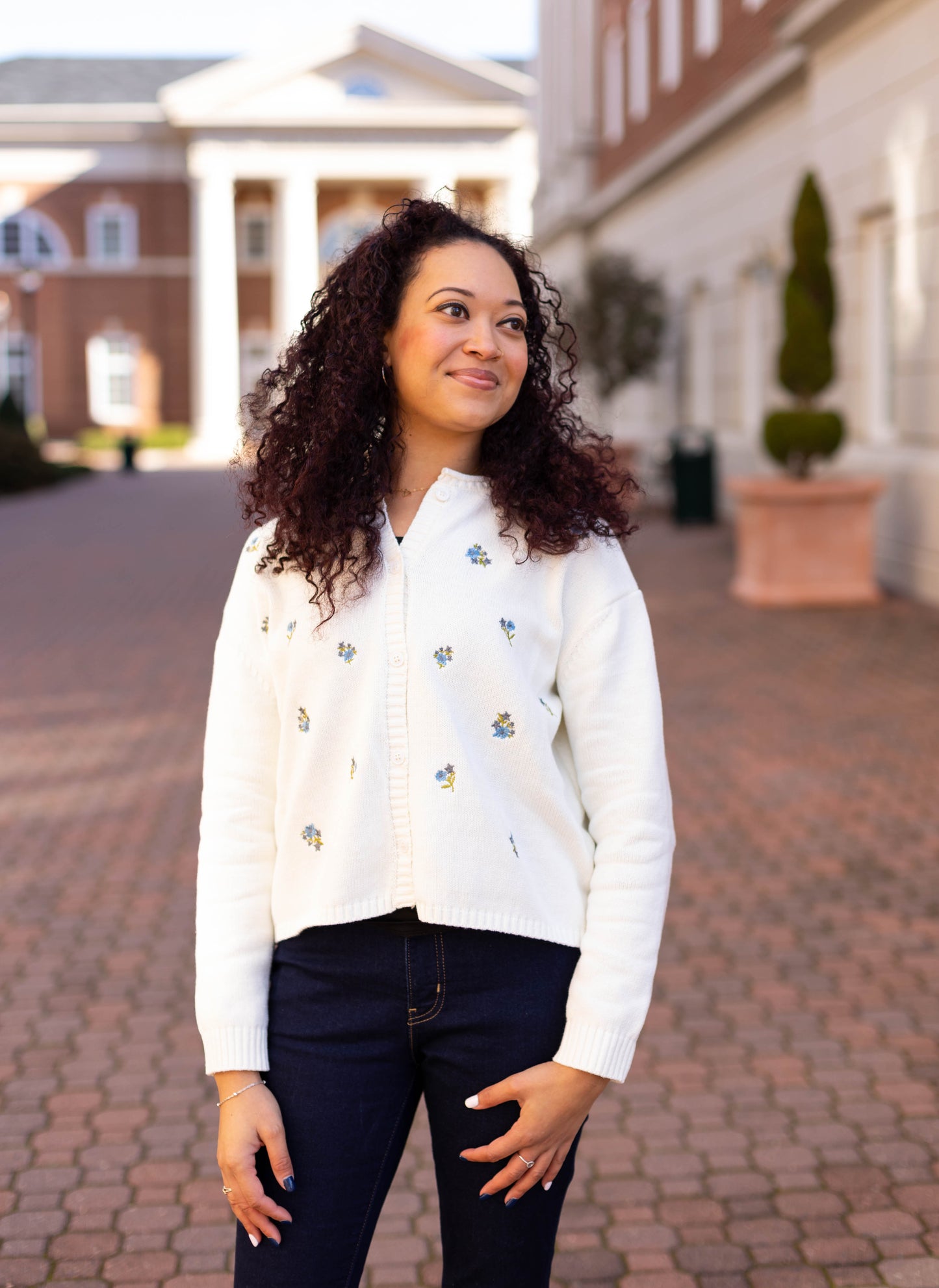 A woman with curly hair smiles on a brick walkway near a building with columns, wearing Dogwood Cloth's Belle Floral Cardigan adorned with embroidered flowers over dark jeans, adding charm to her elegant look.