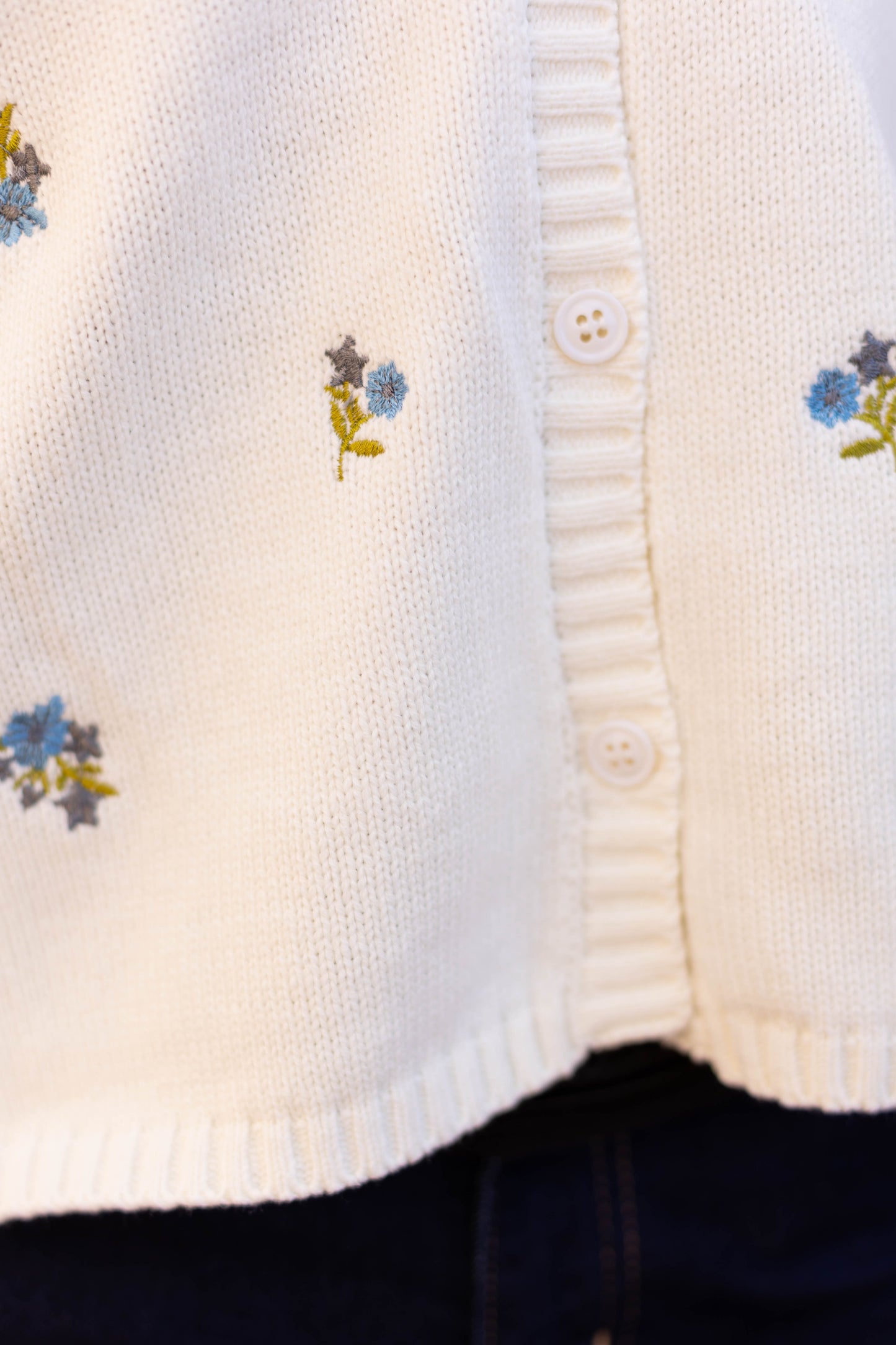 Close-up of Dogwood Cloth's Belle Floral Cardigan, a white knit featuring small embroidered blue and green flowers with white buttons, highlighting its versatility.