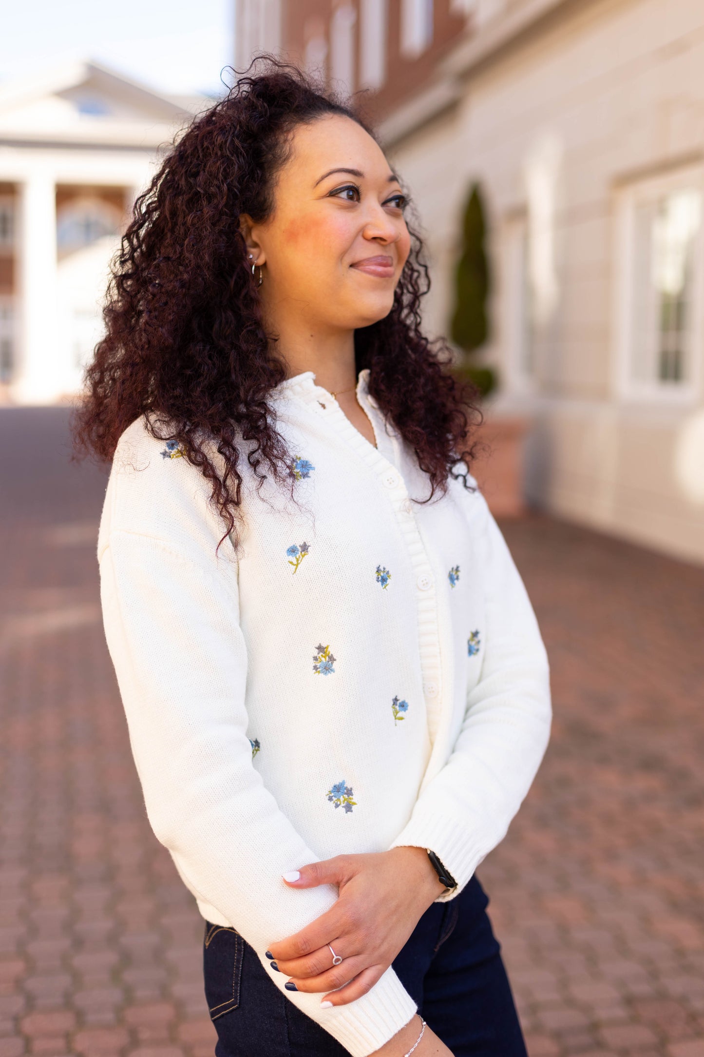 A person with curly hair stands outside a building, smiling slightly with arms crossed while wearing the Dogwood Cloth Belle Floral Cardigan adorned with embroidered flowers.