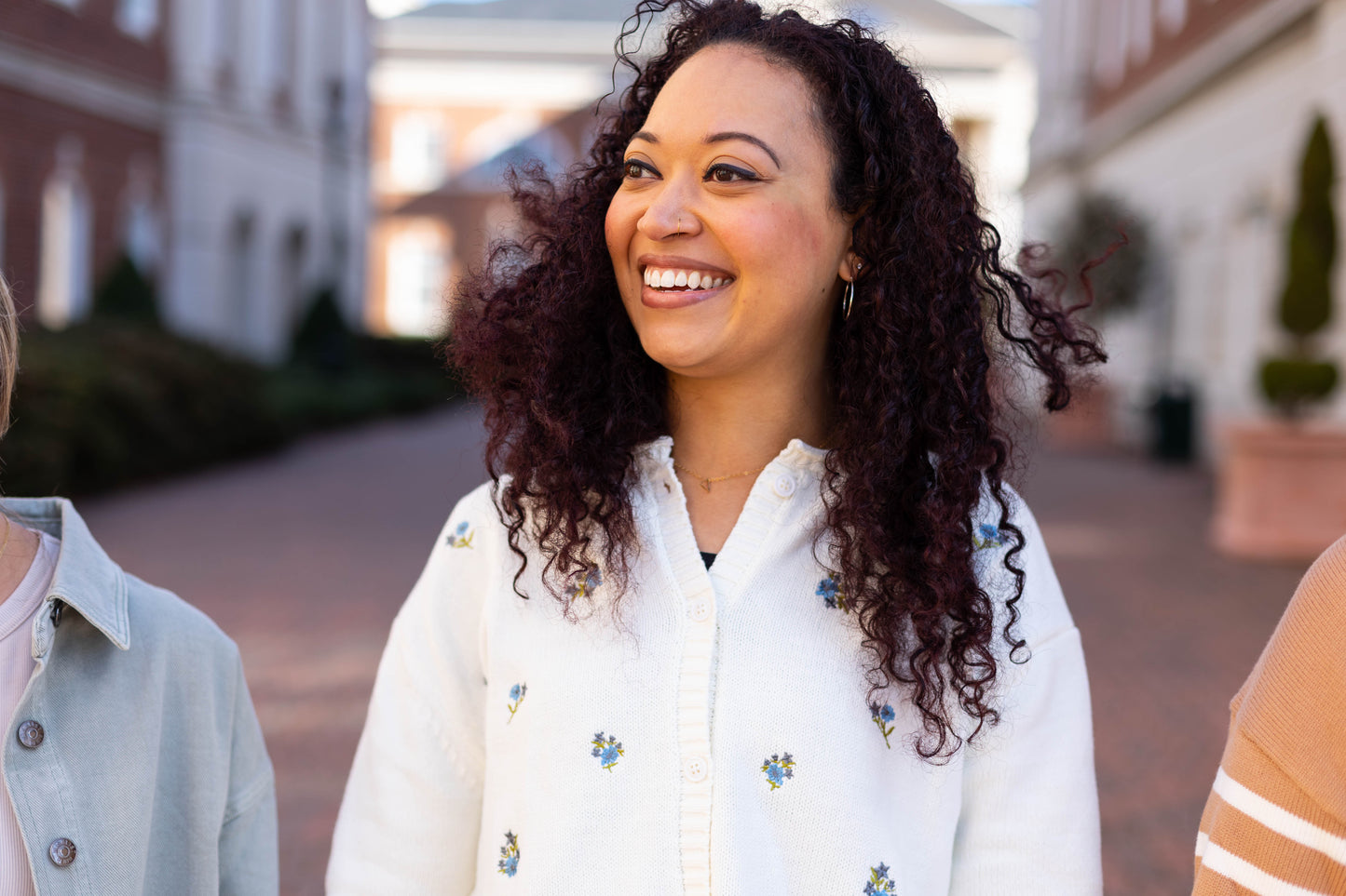 A woman with curly hair smiles outdoors in front of brick buildings, wearing the Belle Floral Cardigan by Dogwood Cloth. The white sweater, adorned with embroidered flowers, perfectly complements her joyful demeanor.