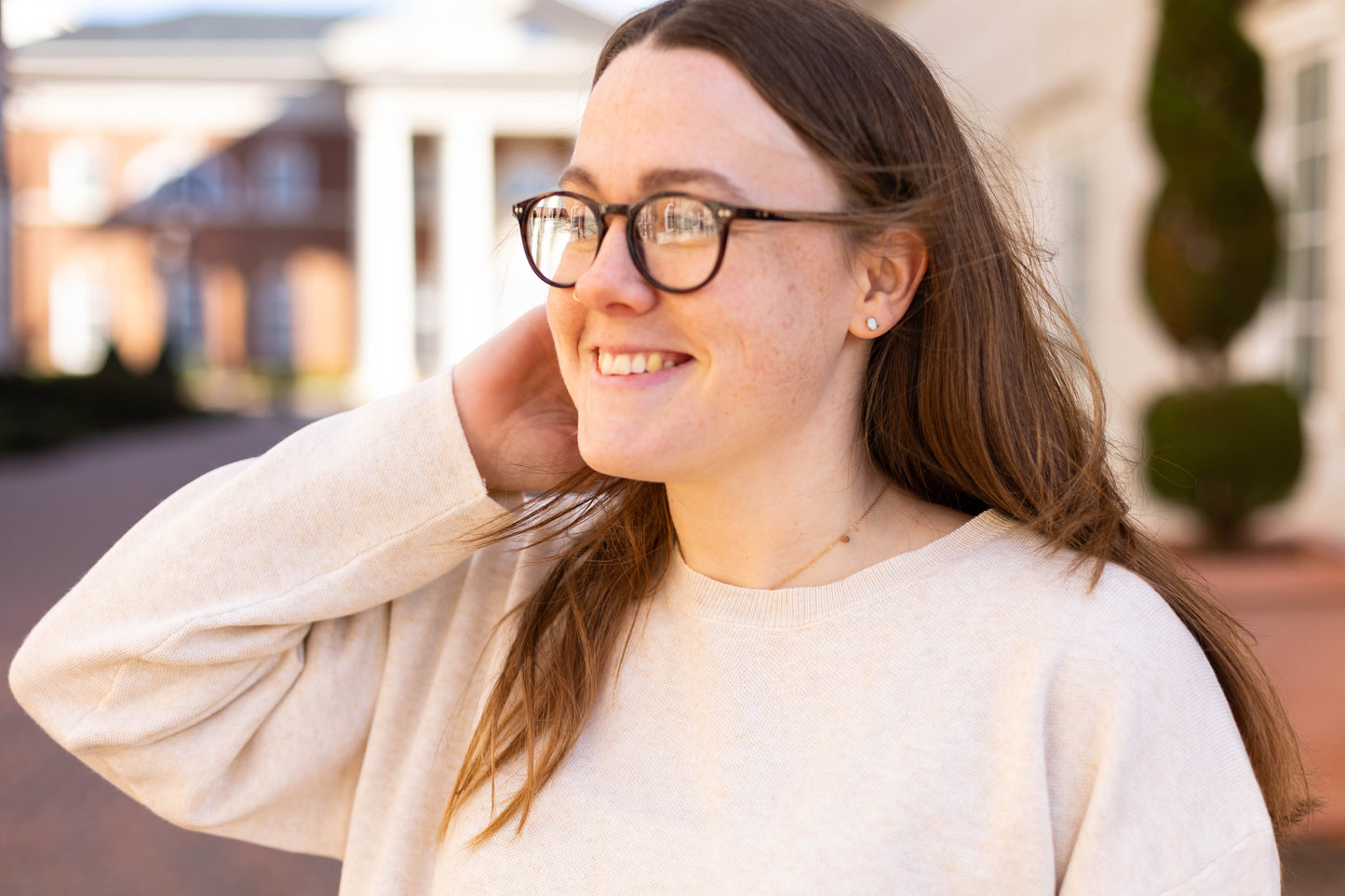 A person with long hair and glasses effortlessly smiles while wearing a Dogwood Cloth Winslet Long-Sleeve sweater, crafted from luxe-feel fabric. Standing near a building with columns outdoors, they exude elegance.