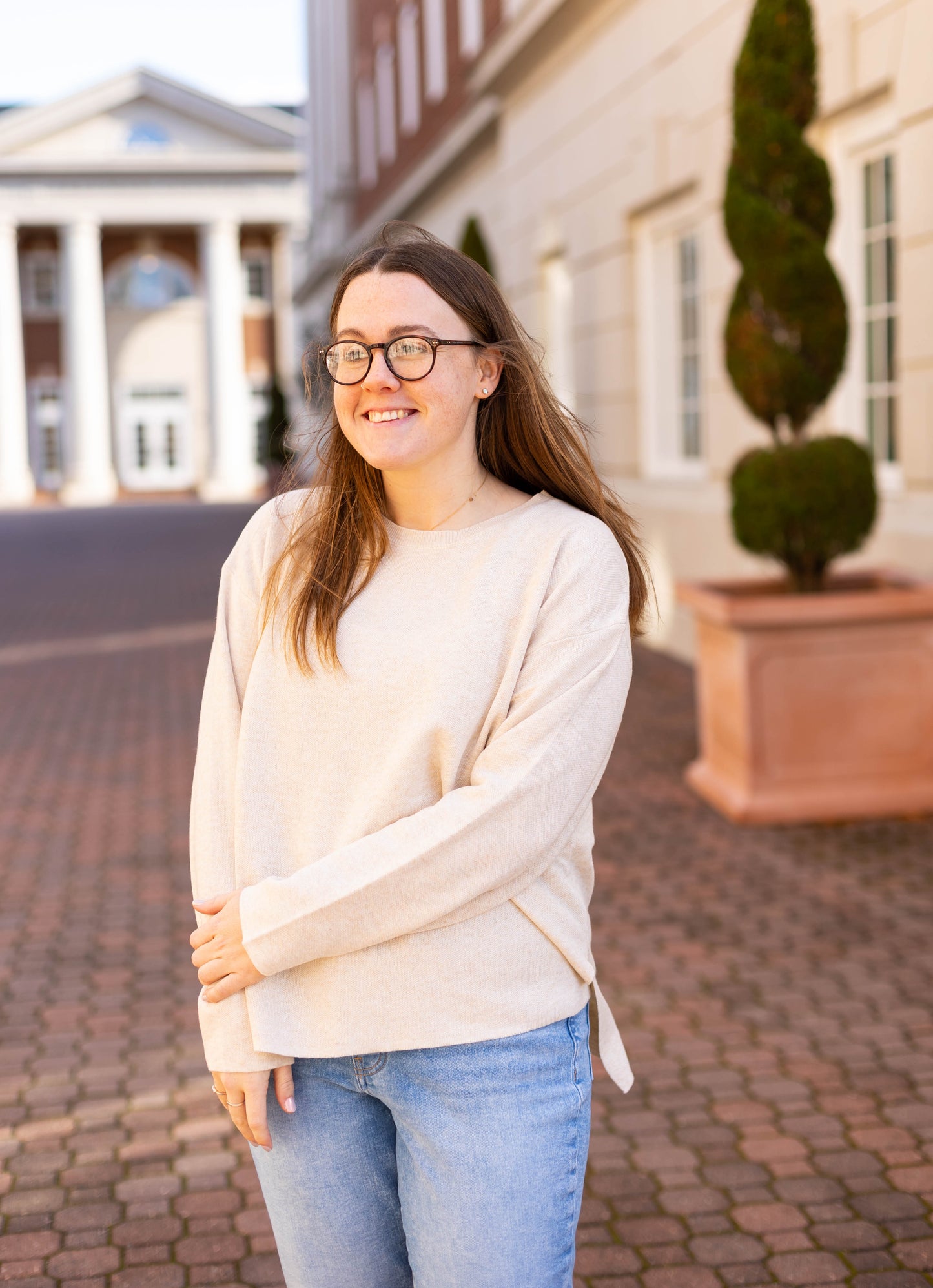 A woman with glasses exudes effortless elegance in a timeless Dogwood Cloth Winslet Long-Sleeve and jeans, standing on a brick pathway in front of a columned building.