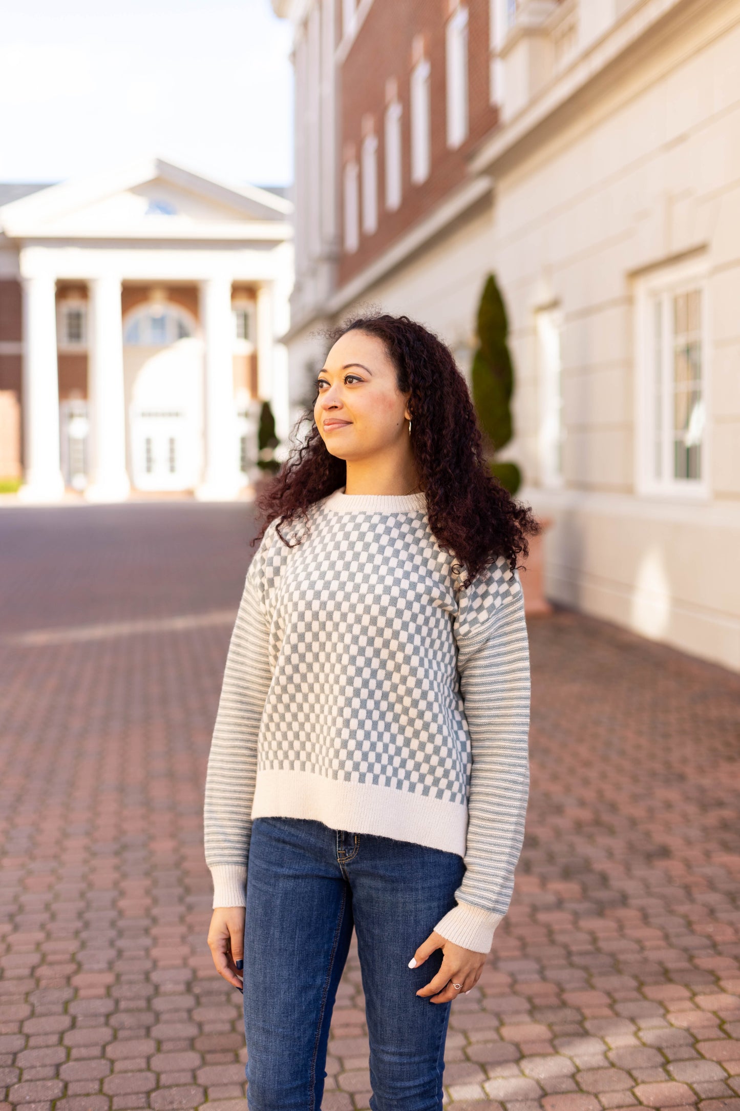 A woman with curly hair in an Emory Contrast Sweater by Dogwood Cloth and jeans stands on a brick pathway, looking to the side, with a building behind her.