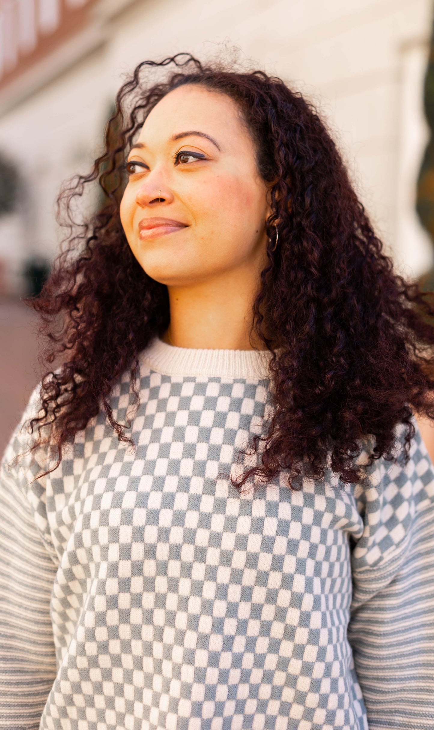 A person with curly hair wearing the Emory Contrast Sweater by Dogwood Cloth looks into the distance with a slight smile while standing outdoors.