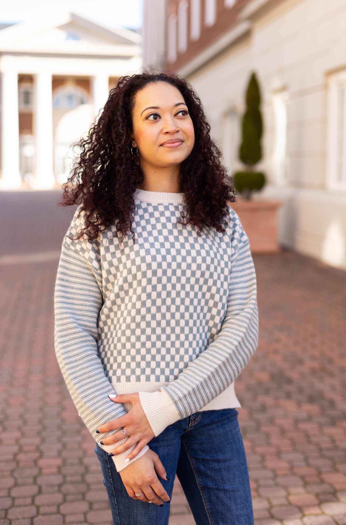 A woman with curly hair stands outside, gazing to the side in her Dogwood Cloth Emory Contrast Sweater and jeans. The cozy chic look perfectly complements the brick path and white building in the background.