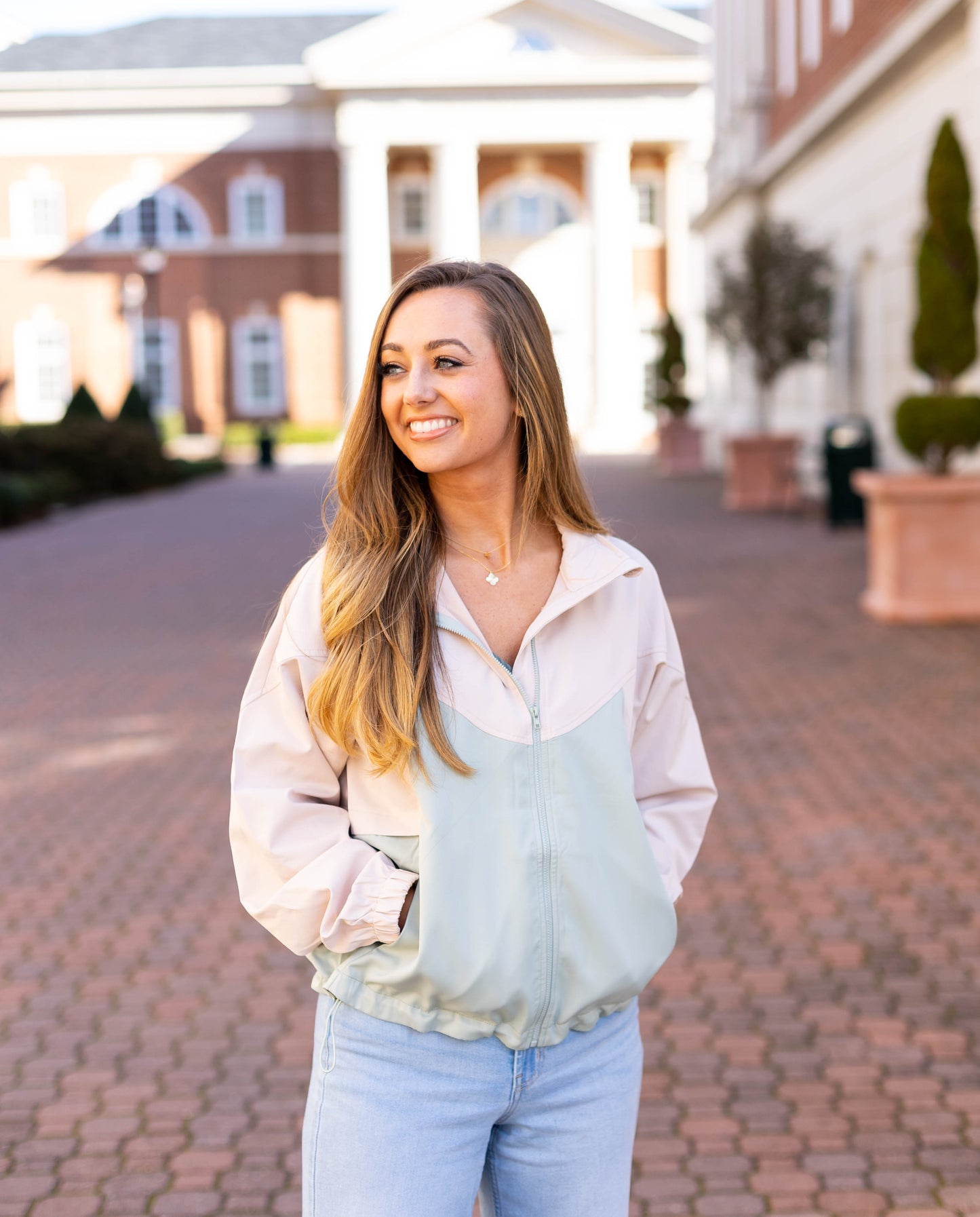 A woman in a Callie Active Jacket by Dogwood Cloth and jeans smiles while standing on a brick pathway with historic buildings in the background.