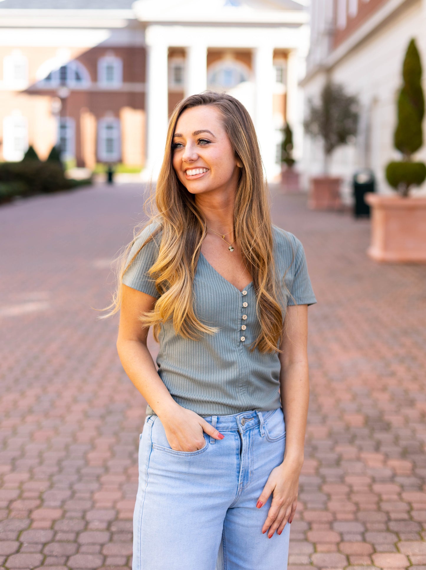Wearing a Dogwood Cloth Greta Everyday Top with a V-neckline and blue jeans, a woman with long hair smiles outdoors on a paved path, buildings in the backdrop.