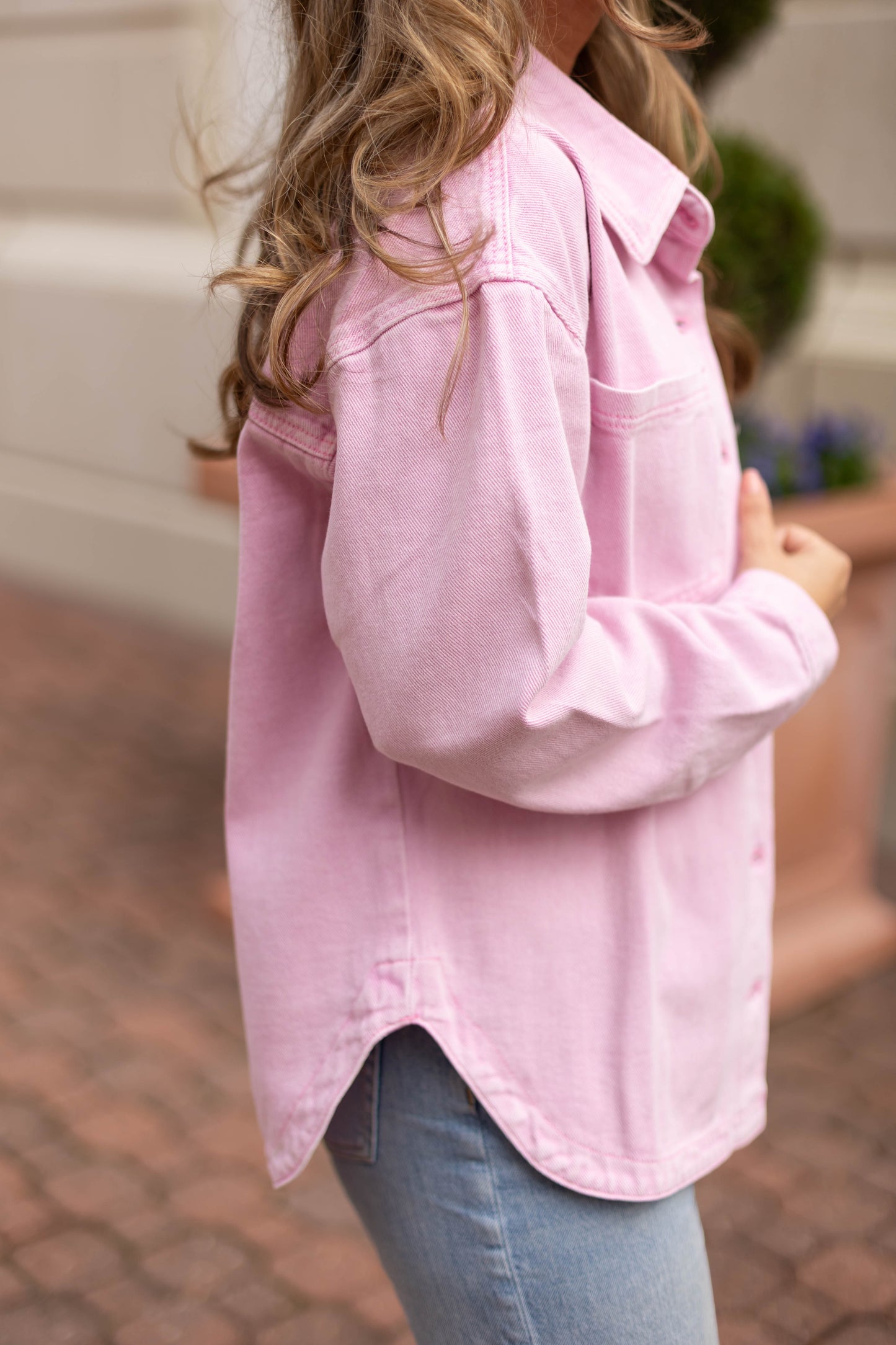 A person with wavy light brown hair, wearing a light pink button-up shirt, light blue jeans, and the Dogwood Cloth Leah Denim Jacket, stands outdoors on a brick walkway.