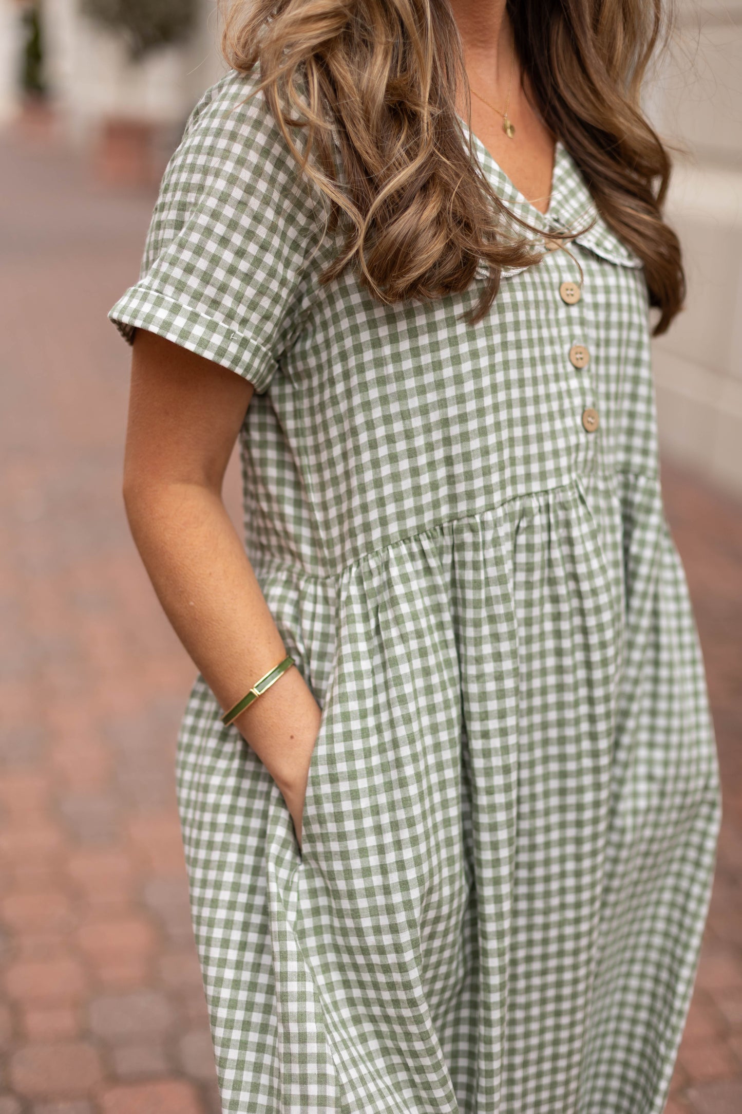 A person stands on a brick walkway, one hand in a pocket, wearing Dogwood Cloth's Caroline Gingham Dress in green and white cotton with buttons and a Peter Pan collar.