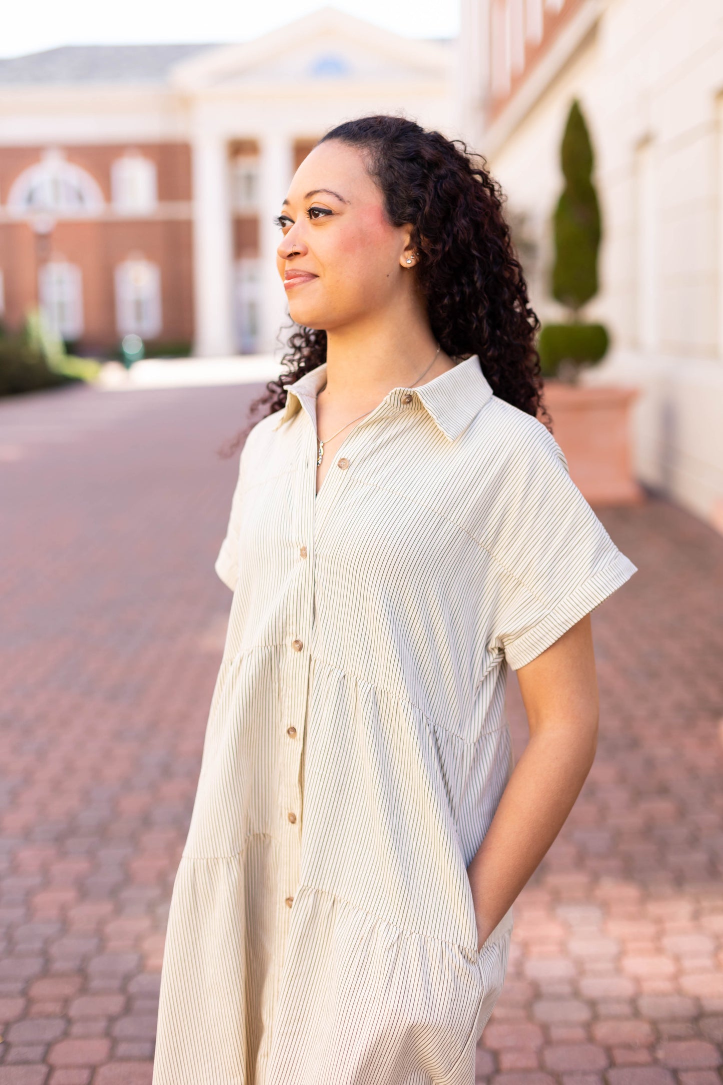 A woman in the Carter Pin-Striped Midi Dress by Dogwood Cloth stands on a brick pathway with her hands in her pockets, gazing to the side. A building with columns is visible in the background.