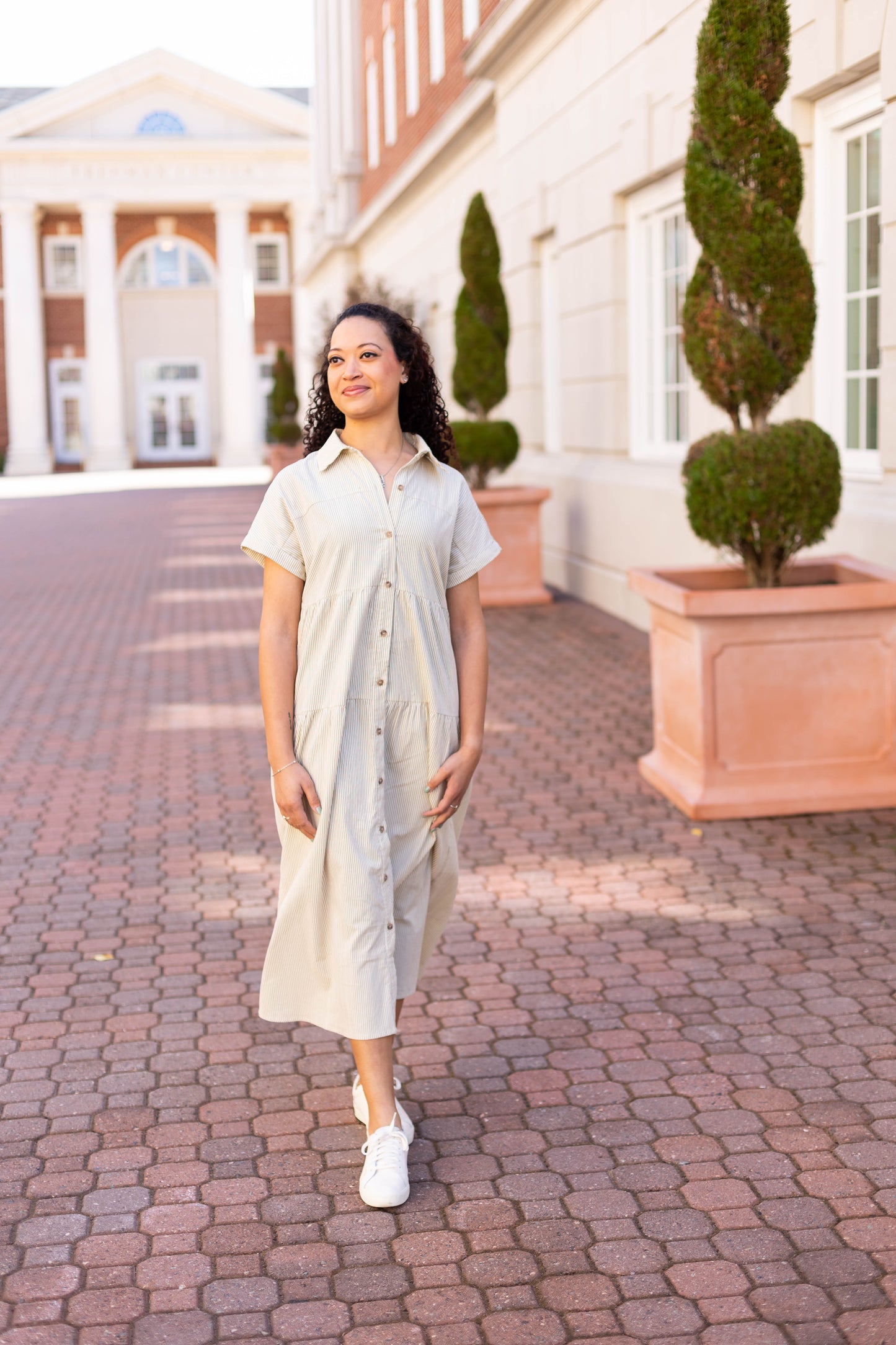 Wearing a Dogwood Cloth Carter Pin-Striped Midi Dress and white sneakers, a person strolls on a brick path amidst trimmed topiary and buildings, with the bright sky overhead.