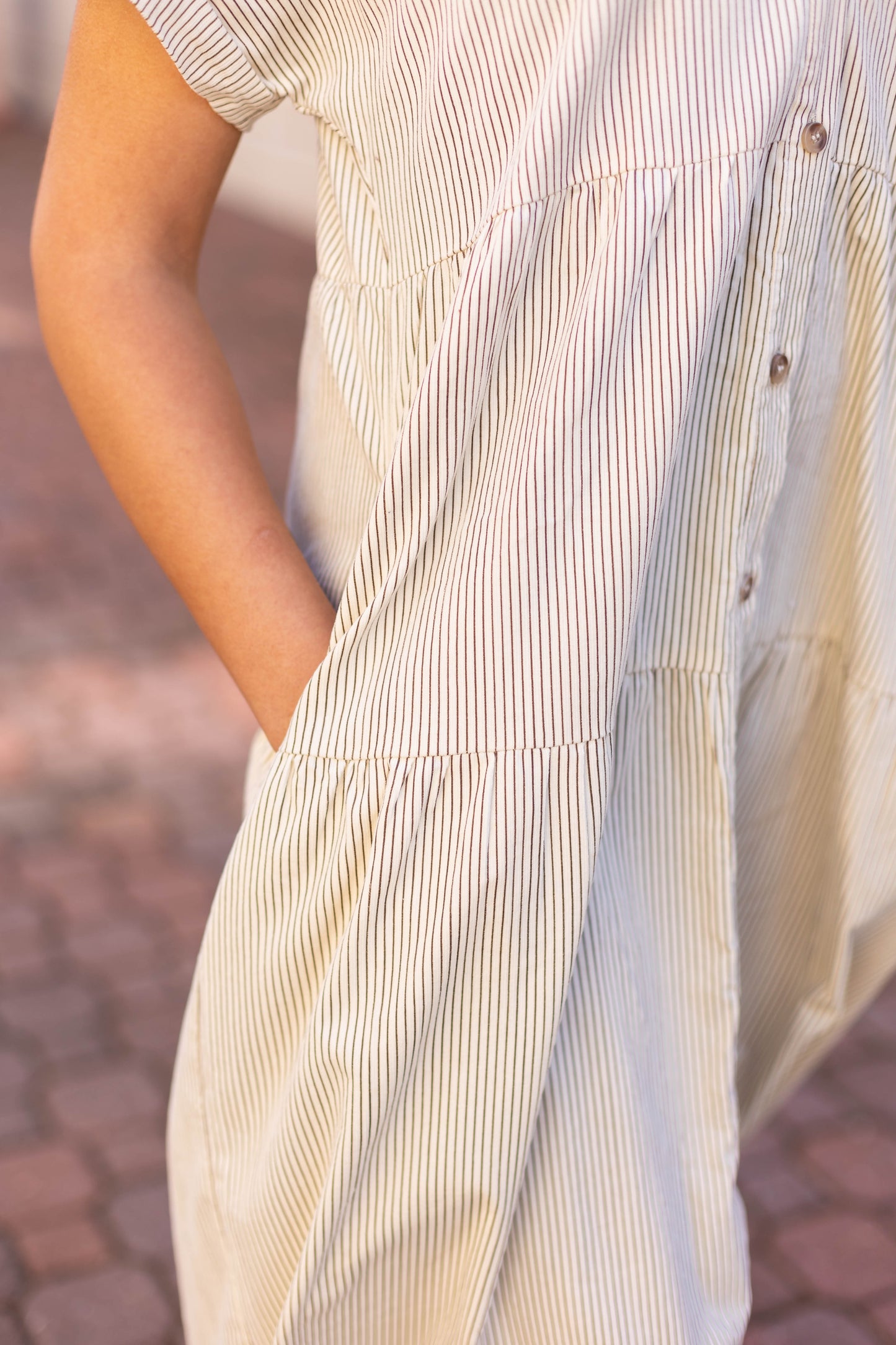 A person stands outdoors in a Carter Pin-Striped Midi Dress by Dogwood Cloth, with short sleeves and one hand in the pocket.