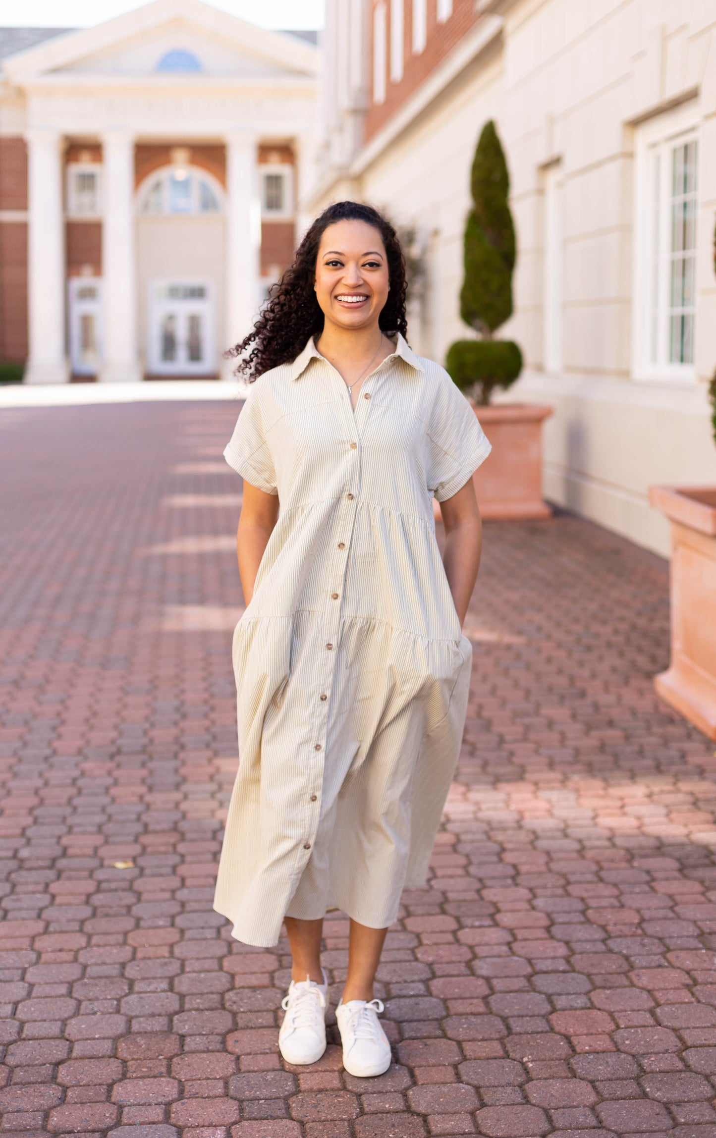 Wearing the Carter Pin-Striped Midi Dress by Dogwood Cloth and sneakers, a person strolls along a paved path between buildings, hands in pockets and smiling brightly.