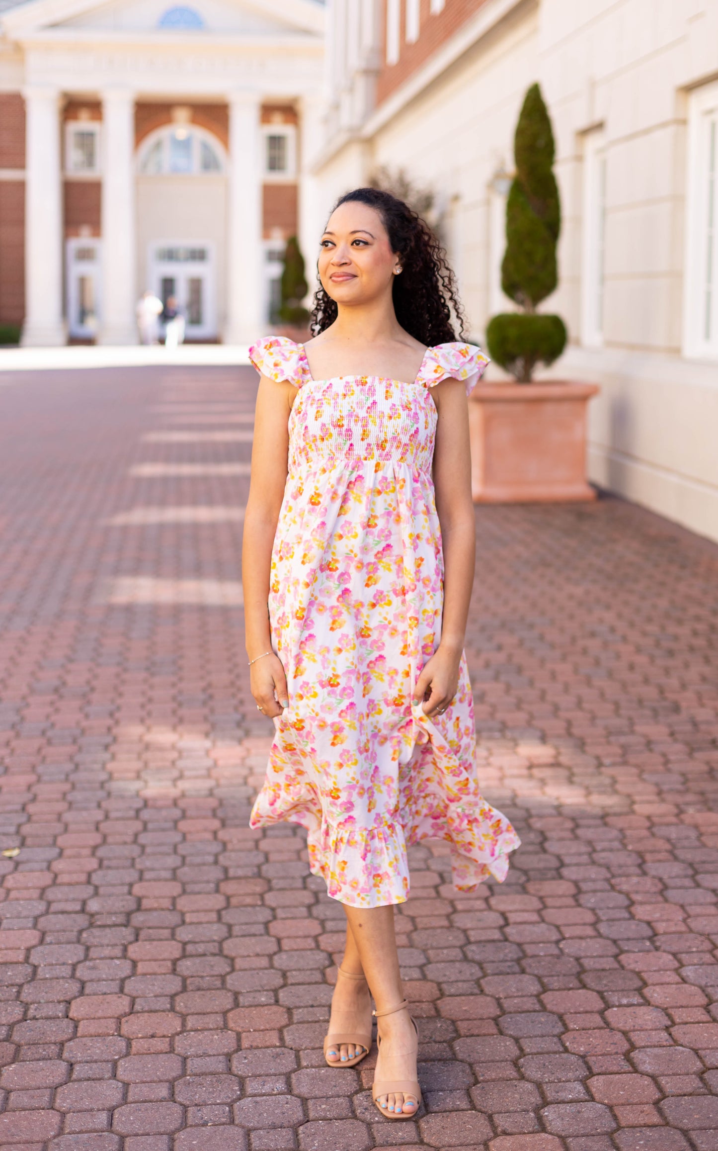 Wearing a Dogwood Cloth Stella Floral Midi with a smocked bust and ruffled hem, a person walks on a brick pathway beside a building adorned with columns and greenery.