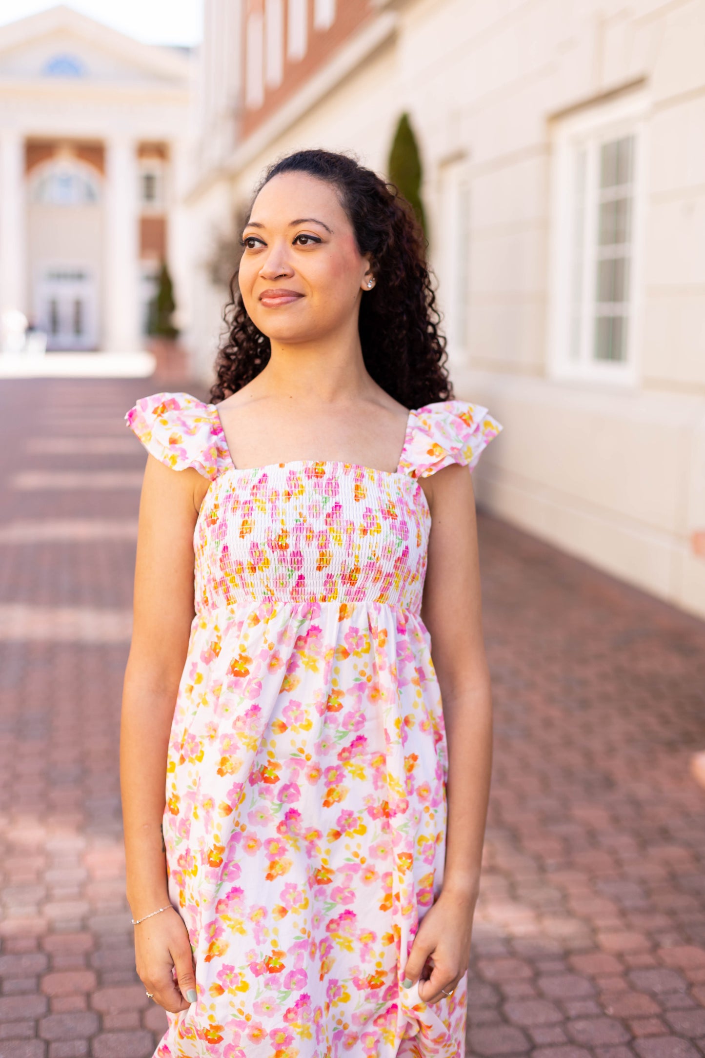 Wearing a Stella Floral Midi by Dogwood Cloth, someone stands on a brick path with a large building in the background, exuding femininity.
