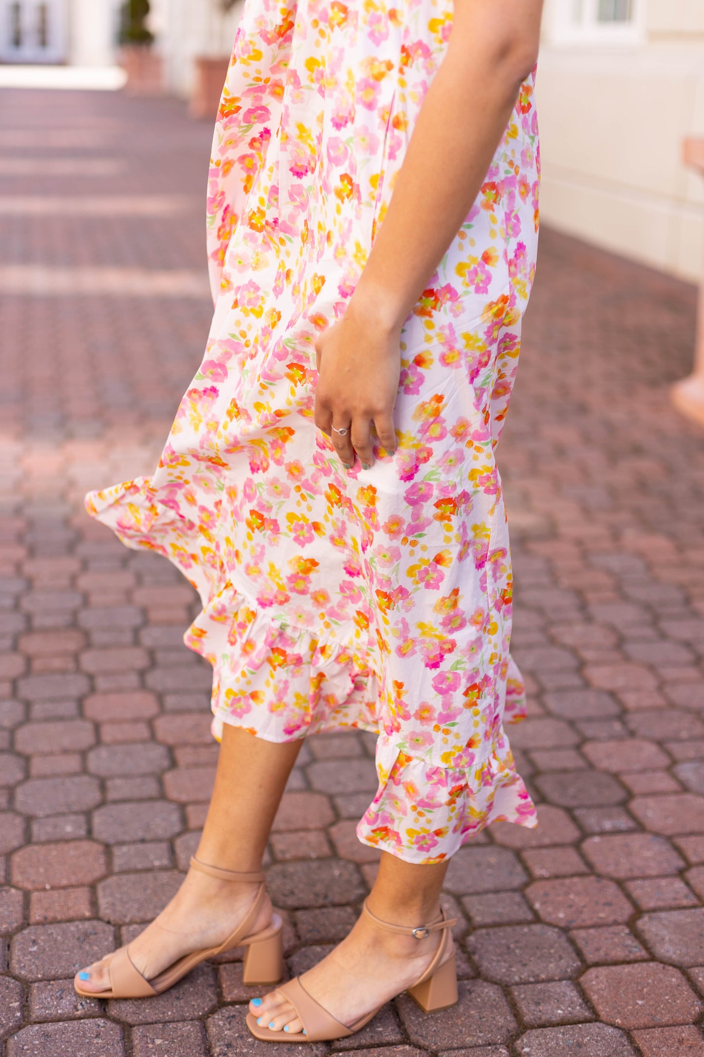 A person in a Stella Floral Midi dress by Dogwood Cloth, featuring a smocked bust and ruffled hem, walks along a brick path in beige sandals.