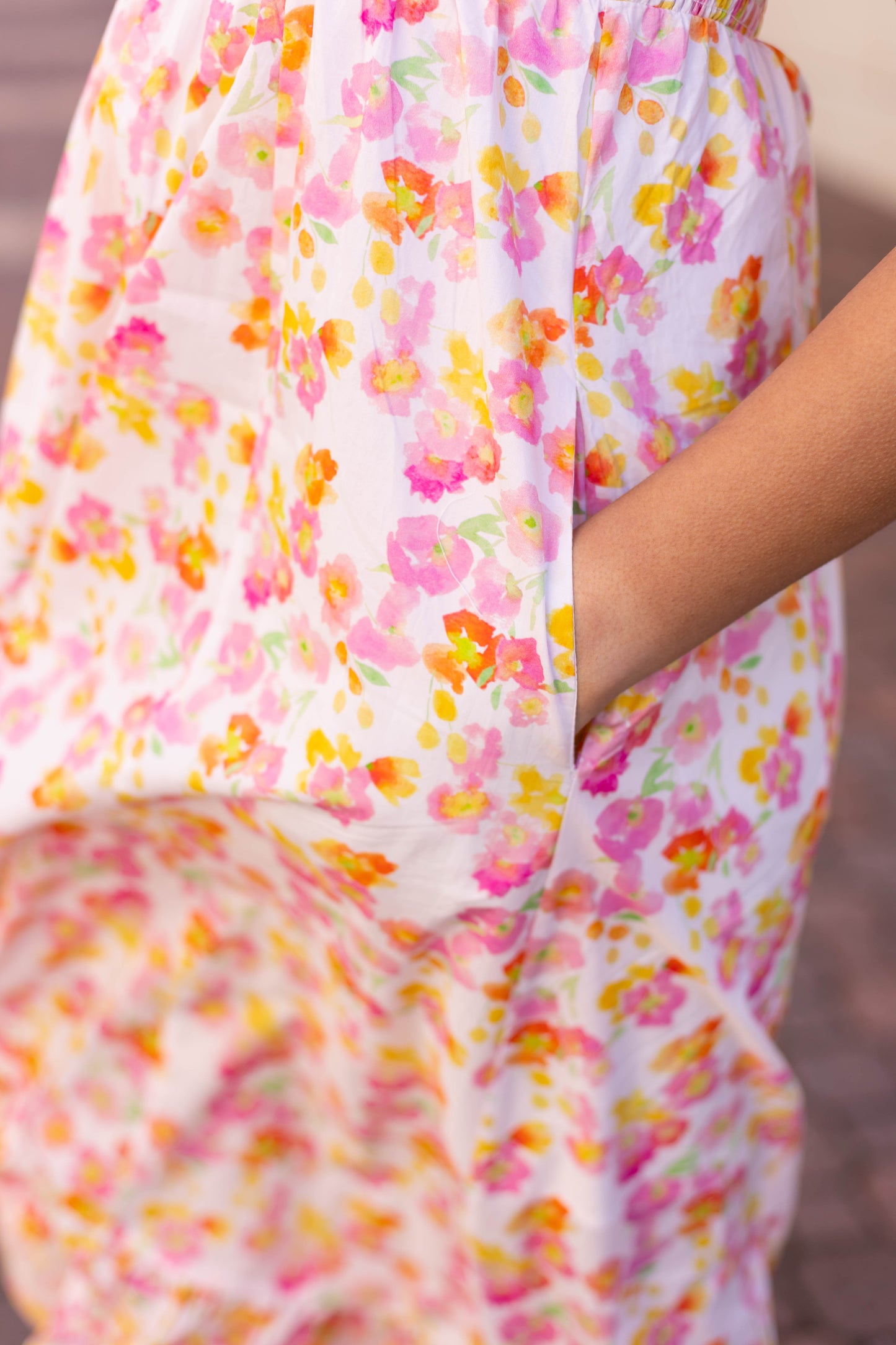 Close-up of someone in the stylish Stella Floral Midi by Dogwood Cloth, showcasing a flowing pink, yellow, and orange floral design. The smocked bust pairs with their hand tucked into a pocket for an elegant look.