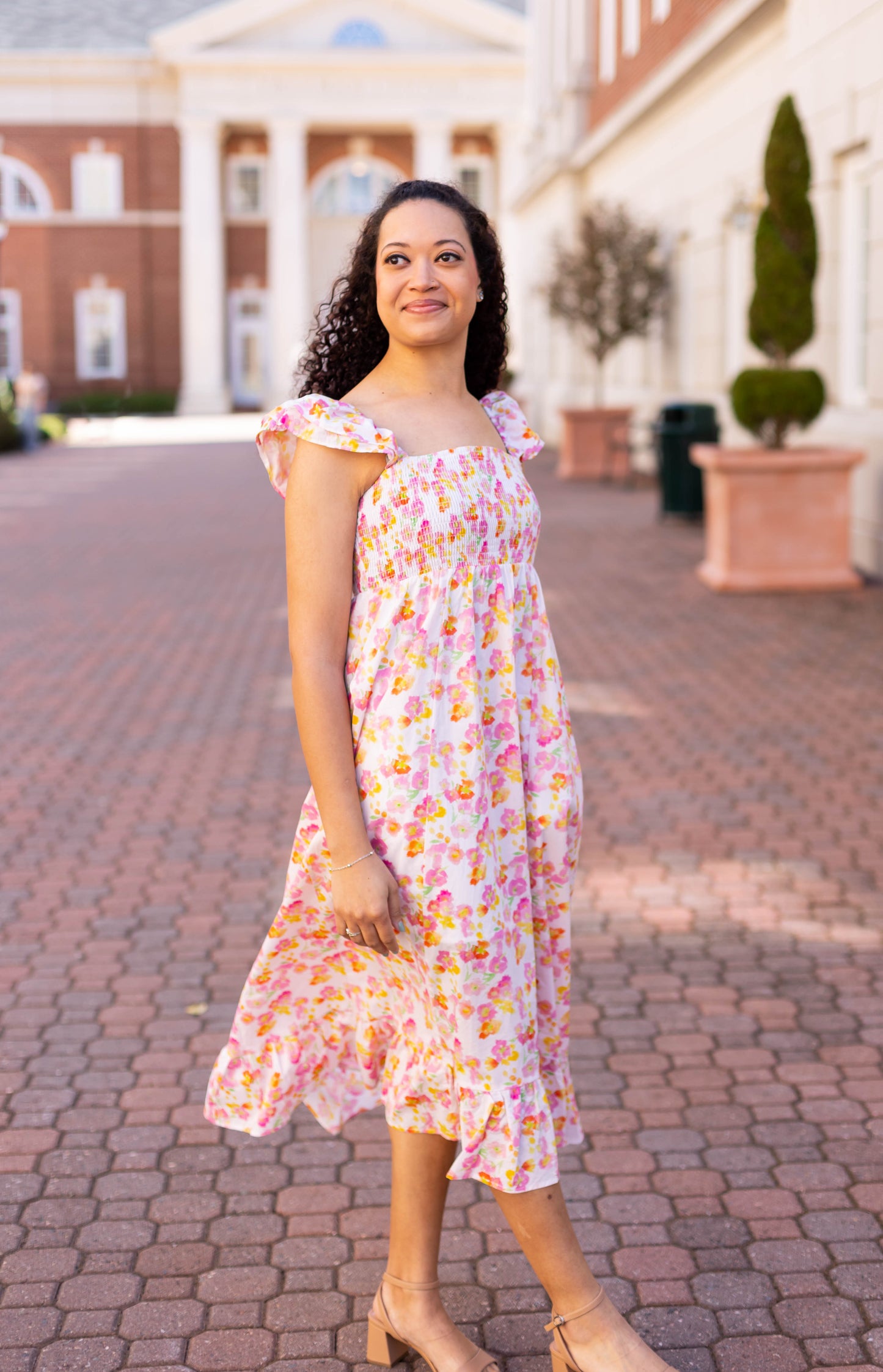 Wearing a Stella Floral Midi by Dogwood Cloth, a person walks gracefully on a brick path, adding femininity against the backdrop of a classical building.