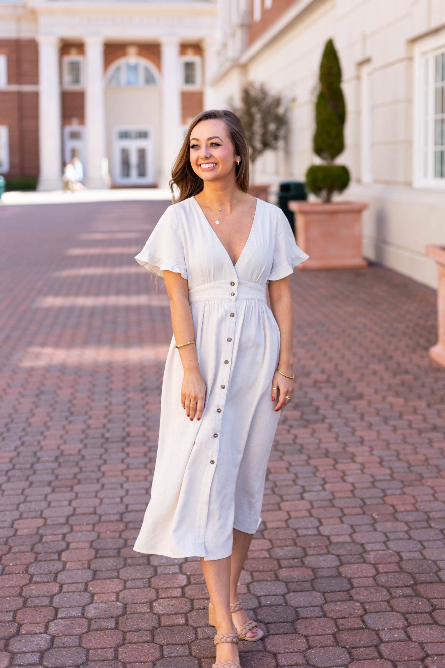 A woman wearing the Dogwood Cloth Chelsea Linen Midi smiles on a brick walkway in front of a grand building with white columns.