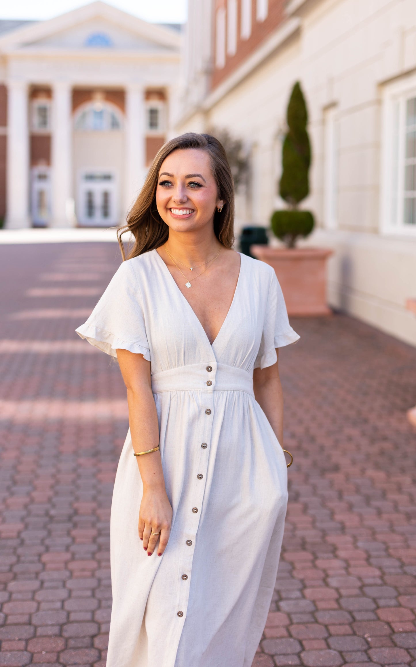 A woman wearing the Dogwood Cloth Chelsea Linen Midi stands smiling on a brick walkway outdoors, with a building featuring columns and large windows in the background.