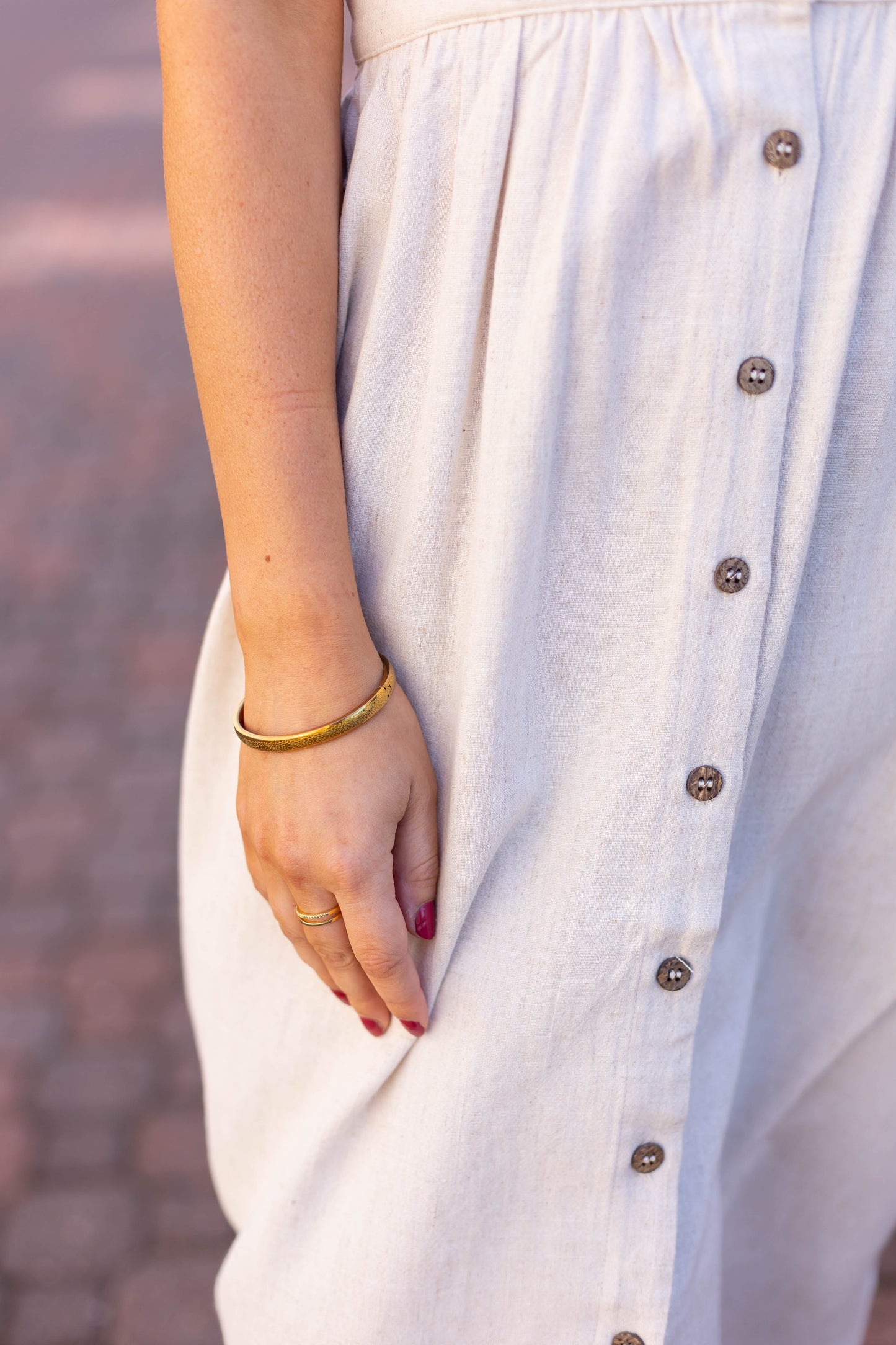 A person in the Dogwood Cloth Chelsea Linen Midi, featuring an A-line silhouette in a light hue, accessorized with a gold bracelet and ring, stands on a paved surface.