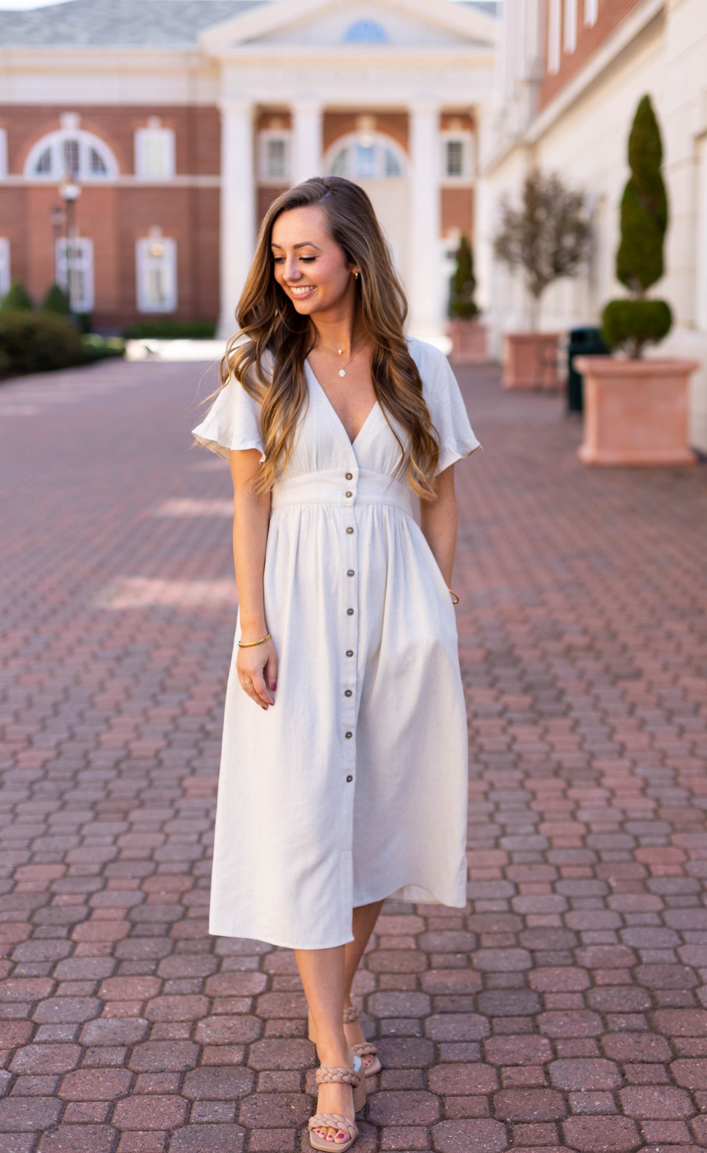 A woman in the Dogwood Cloth Chelsea Linen Midi strolls along a brick pathway before a grand building, embodying the effortless charm of this summer dress.