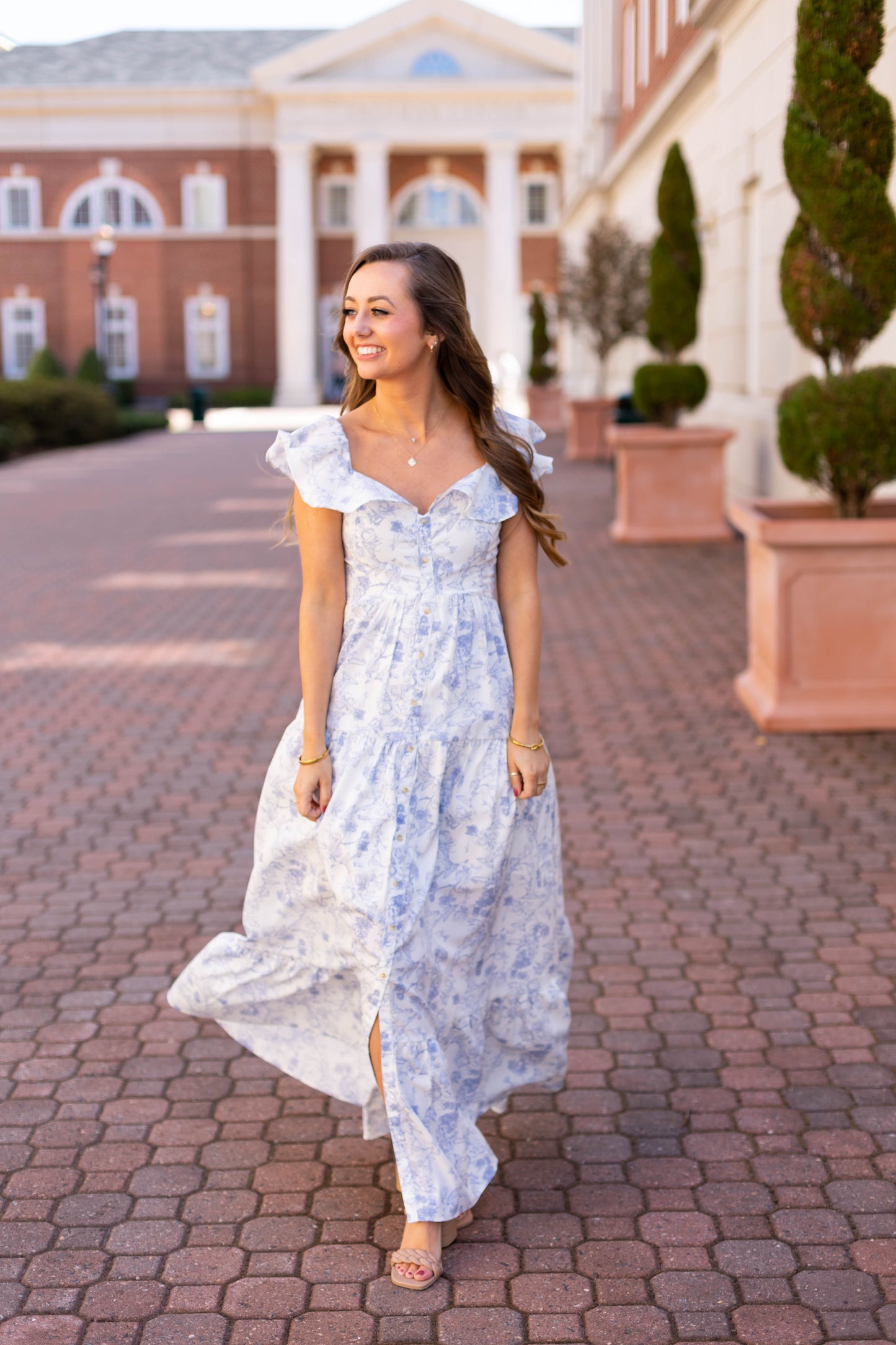 A woman in Dogwood Cloth's Ella Floral Maxi strolls along a brick path, smiling amidst classical-style buildings and topiary. The dress, featuring functional buttons and a charming tiered skirt, complements her graceful surroundings.