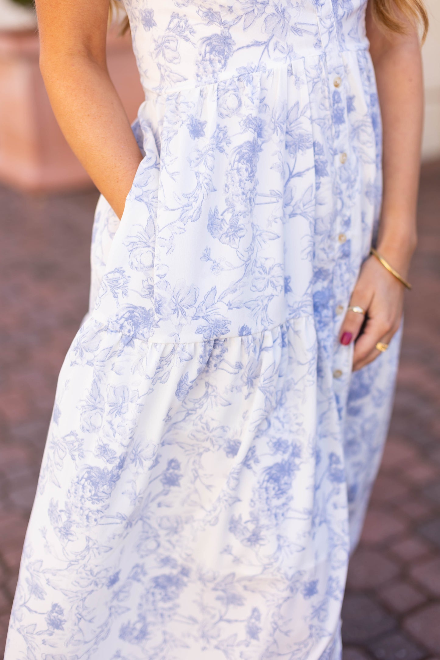 Ella radiates elegance on a cobblestone path, wearing the Dogwood Cloth Ella Floral Maxi—a light blue and white dress with pockets. The functional buttons and tiered skirt enhance her style, perfectly accentuated by a gold bracelet and painted nails.