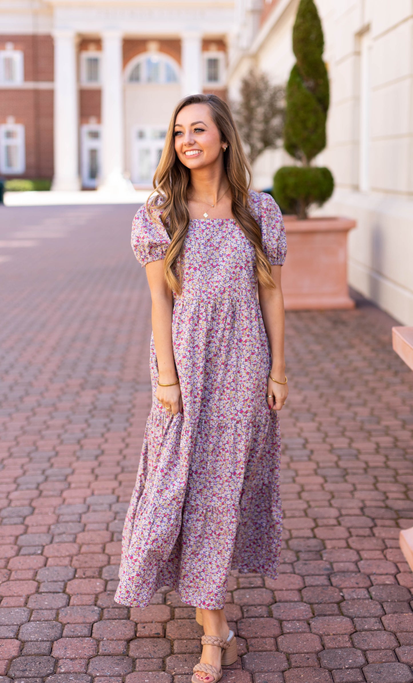 In the breathable cotton of Dogwood Cloth's Jasmine Floral Maxi Dress, a woman exudes effortless elegance as she strolls along a columned brick pathway.
