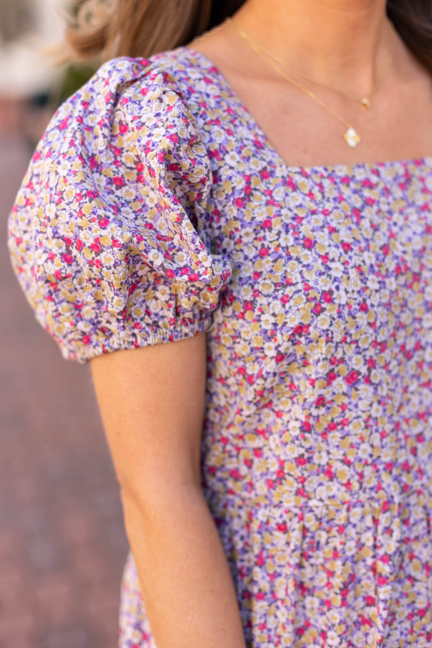 A person dons the Jasmine Floral Maxi Dress by Dogwood Cloth, featuring puffed sleeves and breathable cotton. A gold necklace completes the look against a blurred background, emphasizing the ensemble's effortless elegance.