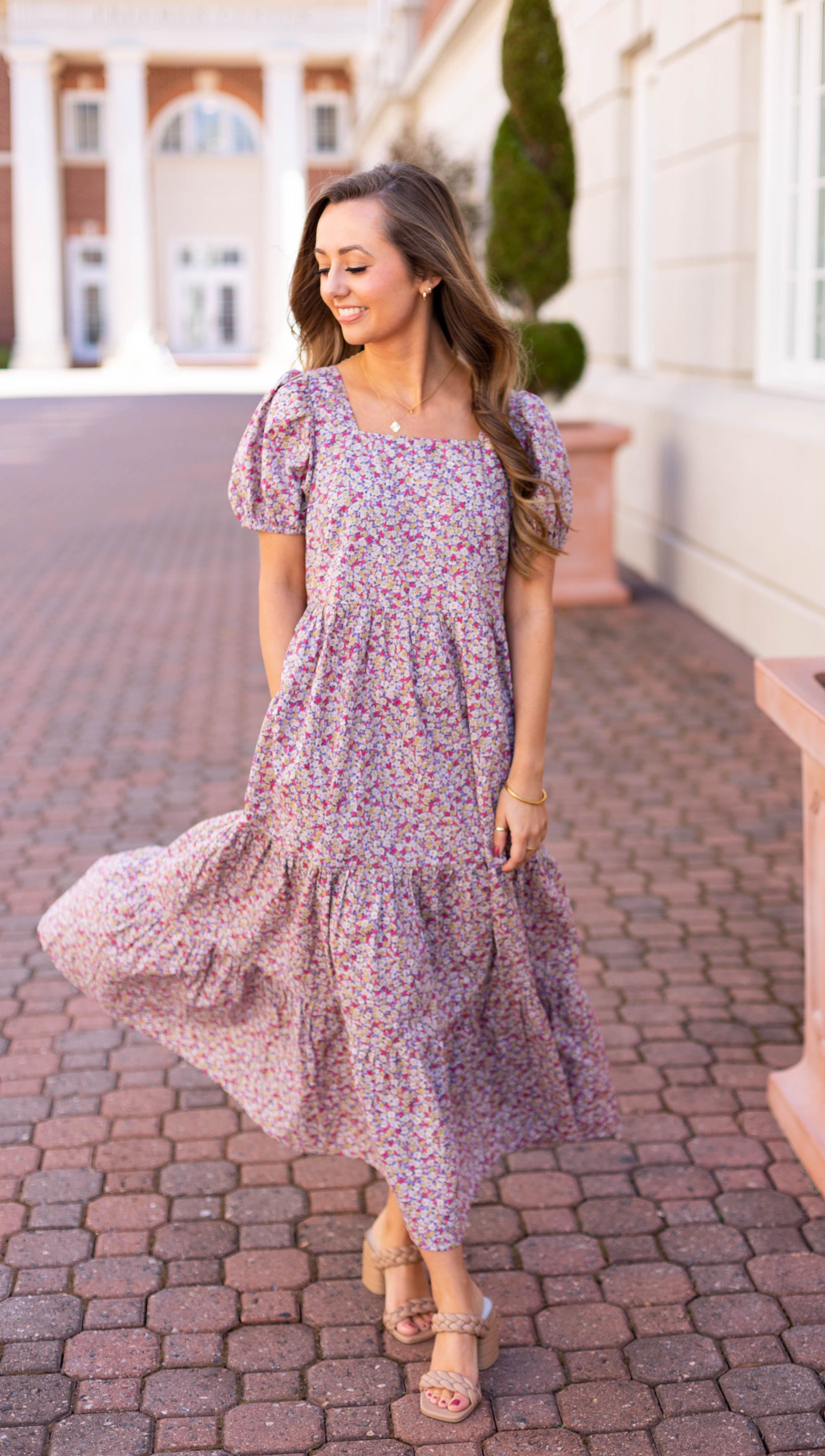 Wearing a Jasmine Floral Maxi Dress by Dogwood Cloth, a woman strolls on a brick path, exuding effortless elegance as she smiles and touches her hair. The breathable cotton fabric catches the breeze, with a column-adorned building standing gracefully in the background.
