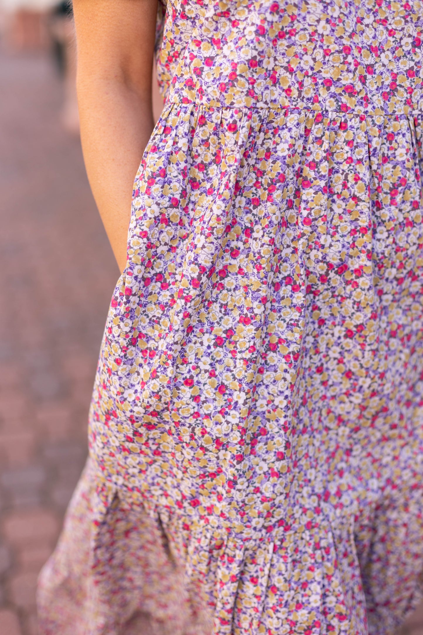 Close-up of a person wearing Dogwood Cloth's Jasmine Floral Maxi Dress in purple, pink, and white. Made from breathable cotton with gathered fabric and visible hemline, exuding elegance as it stands gracefully on a cobblestone path.