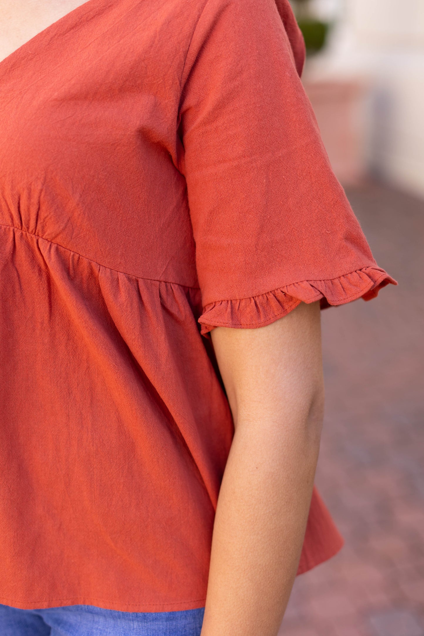 A person stands outside wearing Dogwood Cloth's Jenna Top, featuring a rust-orange babydoll design with a gathered waist.